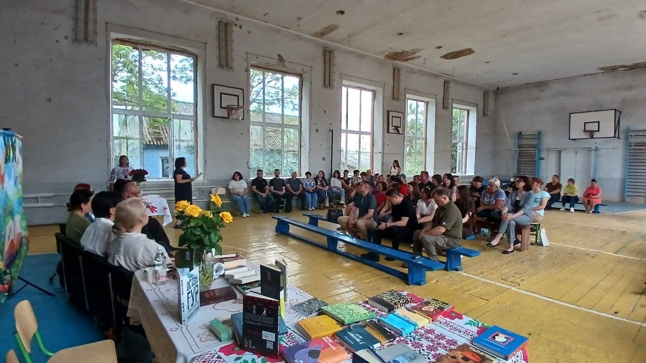 Загальний план великої спортивної зали старої школи, пристосованої під громадську зустріч. По центру кімнати на довгих синіх дерев'яних лавах сидить близько сорока людей різного віку, які слухають виступ. На задньому плані біля великих вікон стоять дві жінки, одна з яких тримає мікрофон. На передньому плані ліворуч облаштовано президію із довгим столом, заваленим книгами, де сидять автори. У правому кутку зали видно баскетбольний щит та гімнастичні лави біля облупленої стіни.