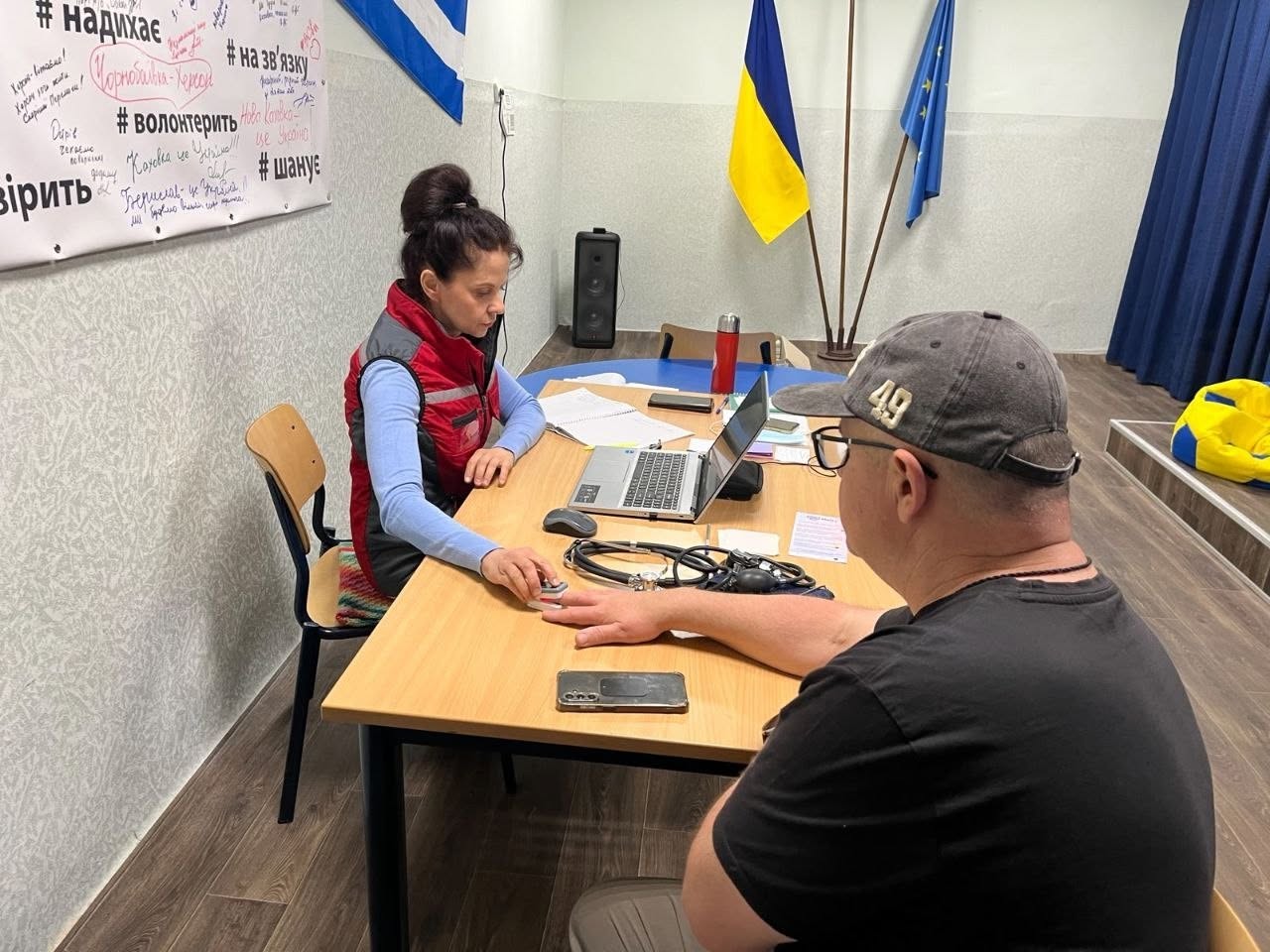 Спеціалістка у червоно-сірому жилеті сидить за робочим столом і проводить медичний огляд відвідувача. Вона тримає руку чоловіка, надіваючи на його вказівний палець пульсоксиметр для вимірювання рівня кисню в крові. Чоловік сидить праворуч боком до камери, одягнений у чорну футболку та сіру кепку з цифрою «49». На столі між ними лежать тонометр, стетоскоп, мобільний телефон, відкритий блокнот і стоїть ноутбук. На задньому плані кімнати встановлено великі прапори України та ЄС біля синьої штори.