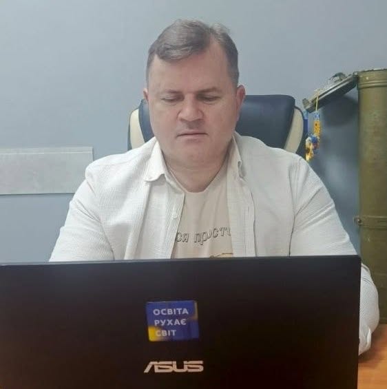 Чоловік середнього віку сидить в офісному кріслі за робочим столом перед екраном ноутбука. Він одягнений у світлу сорочку поверх футболки з частково видимим написом «освіта просто». На передньому плані видно кришку чорного ноутбука «ASUS» із синьо-жовтою наліпкою «ОСВІТА РУХАЄ СВІТ». Праворуч на задньому плані, біля сірої стіни, стоїть гільза від артилерійського снаряда, прикрашена синьо-жовтими стрічками.