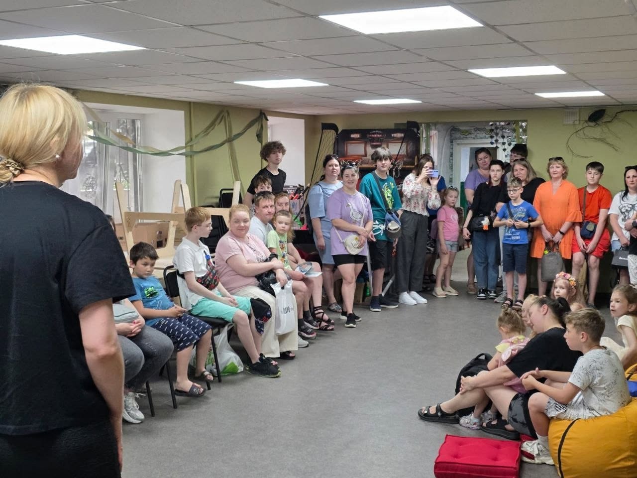 Організаційні збори або лекція для родин у великому світлому залі. На передньому плані ліворуч боком до камери стоїть жінка в чорній футболці, яка звертається до аудиторії. Уздовж лівої стіни на стільцях сидить ряд відвідувачів — переважно жінки та маленькі діти. Решта присутніх (підлітки та дорослі) стоять великою групою біля дальньої стіни. На підлозі праворуч на передньому плані сидить жінка з дітьми на м'яких безкаркасних кріслах-мішках жовтого кольору поруч із червоною квадратною подушкою. Кімната освітлена прямокутними стельовими лампами.