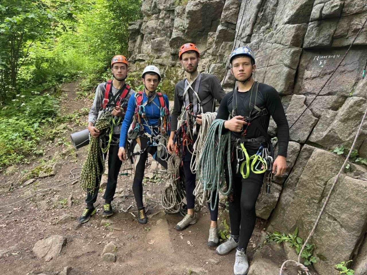 Чотири молоді спортсмени-альпіністи позують перед камерою біля підніжжя високої сірої скелі з кам'яних блоків. Усі хлопці одягнені в спеціальне спорядження: страховочні системи, карабіни, затискачі та захисні каски (дві помаранчеві, одна біла та одна синя). Троє з них тримають у руках великі змотані бухти альпіністських мотузок зеленого та сірого кольорів. На скелі праворуч білою фарбою нанесено маркування маршруту: «Глінтвейн 5Б». Зліва на задньому плані видно ґрунтову лісову стежку.