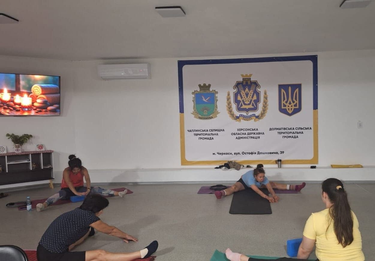 Заняття зі стретчингу або лікувальної фізкультури для жінок у просторій залі хабу. Чотири учасниці різного віку розмістилися на підлозі на індивідуальних спортивних килимках (карематах) і виконують вправу на розтяжку: сидячи з широко розведеними ногами, вони нахиляють тулуб уперед. Дві жінки на передньому плані одягнені у повсякденний спортивний одяг, поруч із ними лежать спеціальні сині блоки для йоги.  На білій стіні по центру кімнати висить великий офіційний банер Чаплинської та Долматівської громад і Херсонської ОВА із зазначенням адреси в Черкасах. Ліворуч на стіні закріплено телевізор, на екрані якого відображається заставка з палаючими свічками та розслаблюючим камінням, а під ним стоїть дерев'яна тумба.