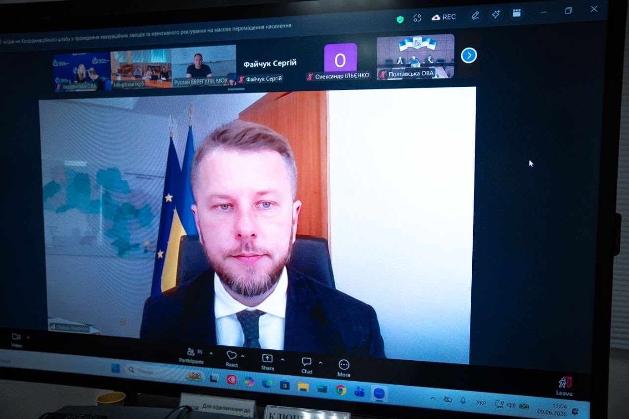 Світлина великого екрана комп'ютера, на якому транслюється засідання Координаційного штабу через сервіс онлайн-нарад. У верхньому лівому кутку видно тему зустрічі: «...засідання Координаційного штабу з проведення евакуаційних заходів та ефективного реагування на масове переміщення населення».  Головне центральне вікно займає трансляція спікера — чоловіка з короткою борідкою у темному піджаку та білій сорочці з краваткою, який виступає на тлі державного прапора України. У верхній стрічці екрана виведені вікна інших учасників, серед яких підписані: «Херсонська ОВА», «Мінрозвитку5», «Руслан БЕРЕГУЛЯ, МОУ», «Файчук Сергій», «Олександр ІЛЬЄНКО» та «Полтавська ОВА». У правому нижньому кутку панелі завдань Windows відображається дата «09.06.2026» та час «13:04».
