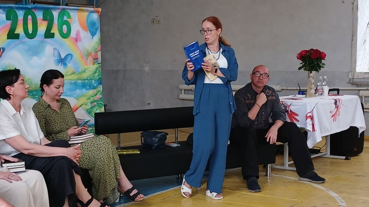 Виступ письменниці або спікерки перед авдиторією в залі. У центрі кадру стоїть рудоволоса жінка в окулярах та синьому костюмі (брюки й вільна сорочка поверх білого топа), яка тримає в руках синю книгу та папери, презентуючи видання. Праворуч від неї на чорній кушетці сидить чоловік в окулярах та темній вишиванці, уважно слухаючи виступ. Ліворуч на стільцях сидять дві жінки: одна в білій сорочці, інша — в зеленій сукні з дрібним візерунком. Позаду них встановлено яскравий дитячий банер із великим написом «2026», веселкою та метеликами.