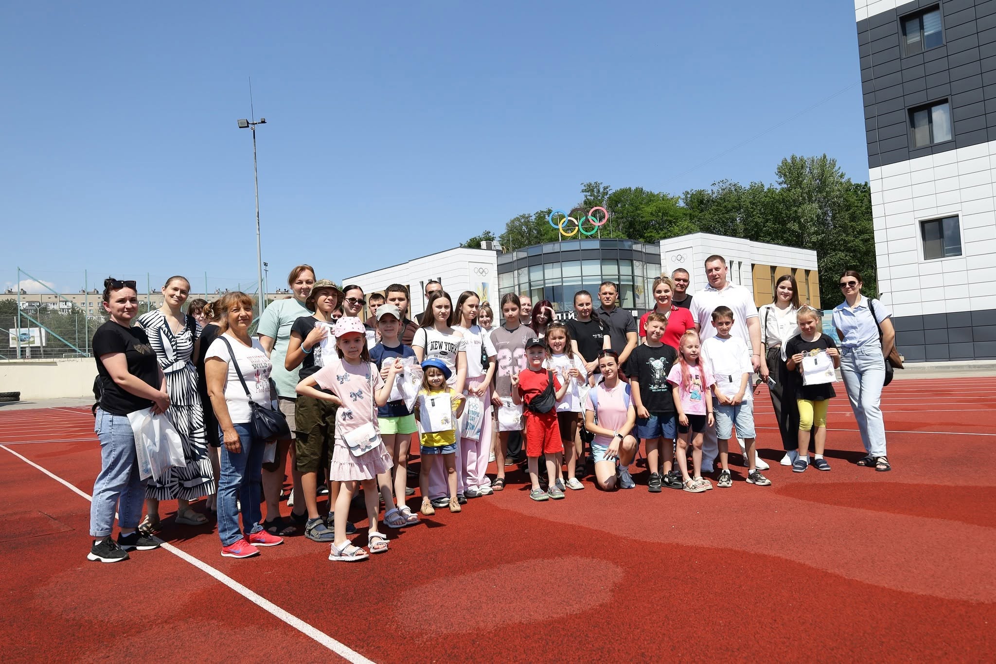 Велике групове фото учасників заходу на відкритому спортмайданчику з червоним гумовим покриттям. Близько тридцяти людей — діти різного віку та дорослі — вишикувалися в один ряд під яскравим безхмарним небом. Більшість одягнені в літній повсякденний або спортивний одяг, дехто з дітей тримає в руках білі подарункові пакети. На задньому плані по центру видно сучасну будівлю з панорамним склінням, на даху якої встановлено об'ємні різнокольорові олімпійські кільця. Праворуч височіє багатоповерхова офісна будівля зі світлим облицюванням фасаду.