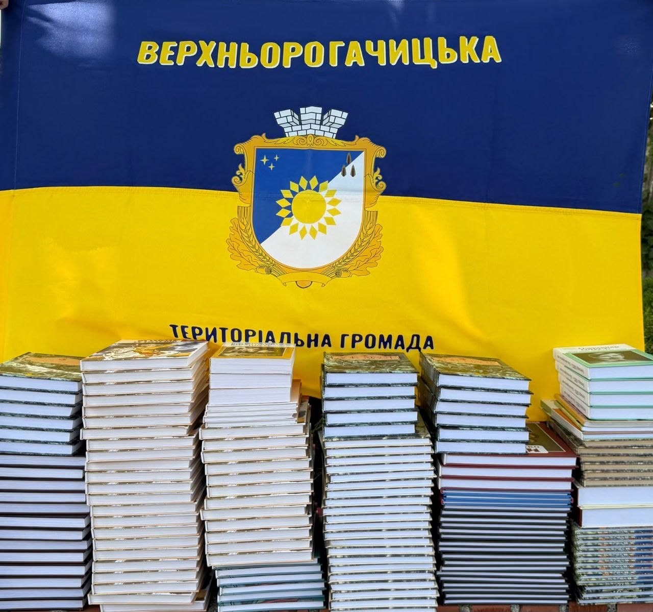 Великі рівні стопки нових книг у твердих та м'яких обкладинках вишикувані в ряд на тлі офіційного прапора громади. Прапор розділений горизонтально на дві рівні частини: верхню синю та нижню жовту. У центрі полотнища зображено герб із соняшником та міською короною у формі мурованої вежі. У верхній частині прапора нанесено великий напис жовтими літерами: «ВЕРХНЬОРОГАТИЧСЬКА», а внизу на межі кольорів чорним шрифтом написано: «ТЕРИТОРІАЛЬНА ГРОМАДА». Перед прапором стоять шість щільних вертикальних пачок із книжками різної товщини.