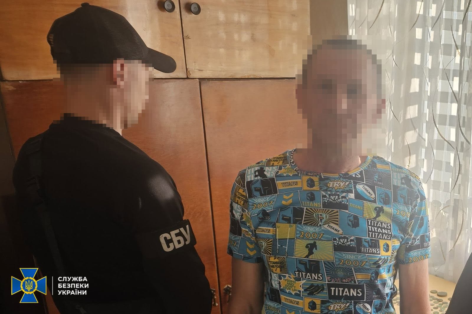 Співробітник СБУ стоїть поруч із затриманим у кімнаті біля дерев'яної стіни-меблів. Ліворуч у напівпрофіль зображено оперативника у чорній тактичній кепці, чорній кофті з білим написом «СБУ» на рукаві та з наплічною чорною сумкою. Його обличчя повністю приховане піксельним розмиттям.  Праворуч стоїть чоловік середнього віку у яскравій різнокольоровій футболці з принтом у вигляді прямокутних синьо-жовтих комікс-блоків із написами «TITANS», «SINCE 2007» та «TRY!». Обличчя затриманого також повністю розмите цифровим фільтром.