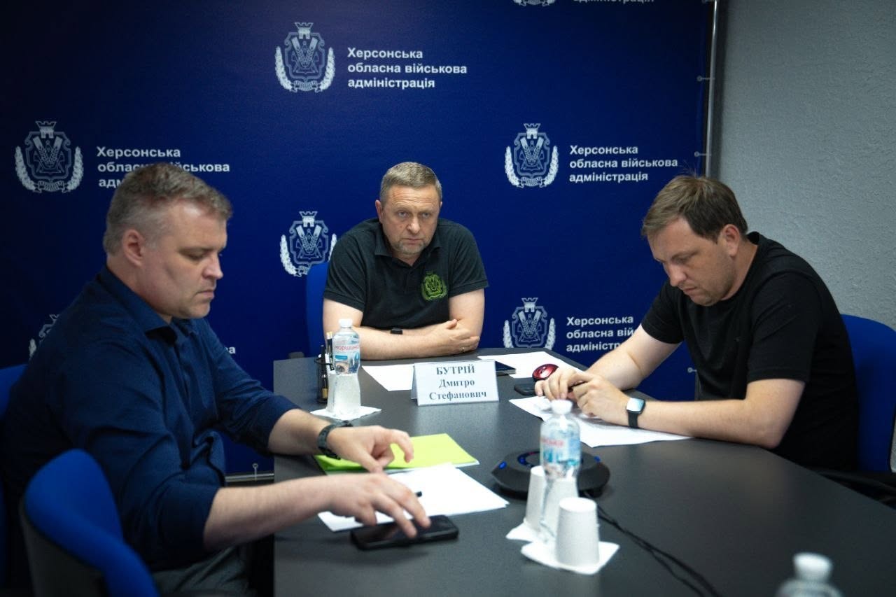 Троє представників керівництва Херсонської ОВА беруть участь у засіданні за довгим робочим столом сірого кольору. По центру сидітиме Дмитро Бутрій у чорній футболці-поло, перед яким стоїть іменна табличка «БУТРІЙ Дмитро Стефанович». Ліворуч від нього сидить чоловік у темно-синій сорочці, який схилився над паперами й тримає в руках мобільний телефон. Праворуч за столом сидить інший чоловік у чорній футболці, який робить нотатки ручкою на аркуші паперу.  На столі перед учасниками стоять пластикові пляшки з водою, паперові стаканчики та розміщено пристрій гучного зв'язку (спікерфон). Тло за спинами посадовців оформлене темно-синім пресволлом із написом «Херсонська обласна військова адміністрація» та білими гербами області. Права частина кадру за столом межує із сірою стіною кабінету.