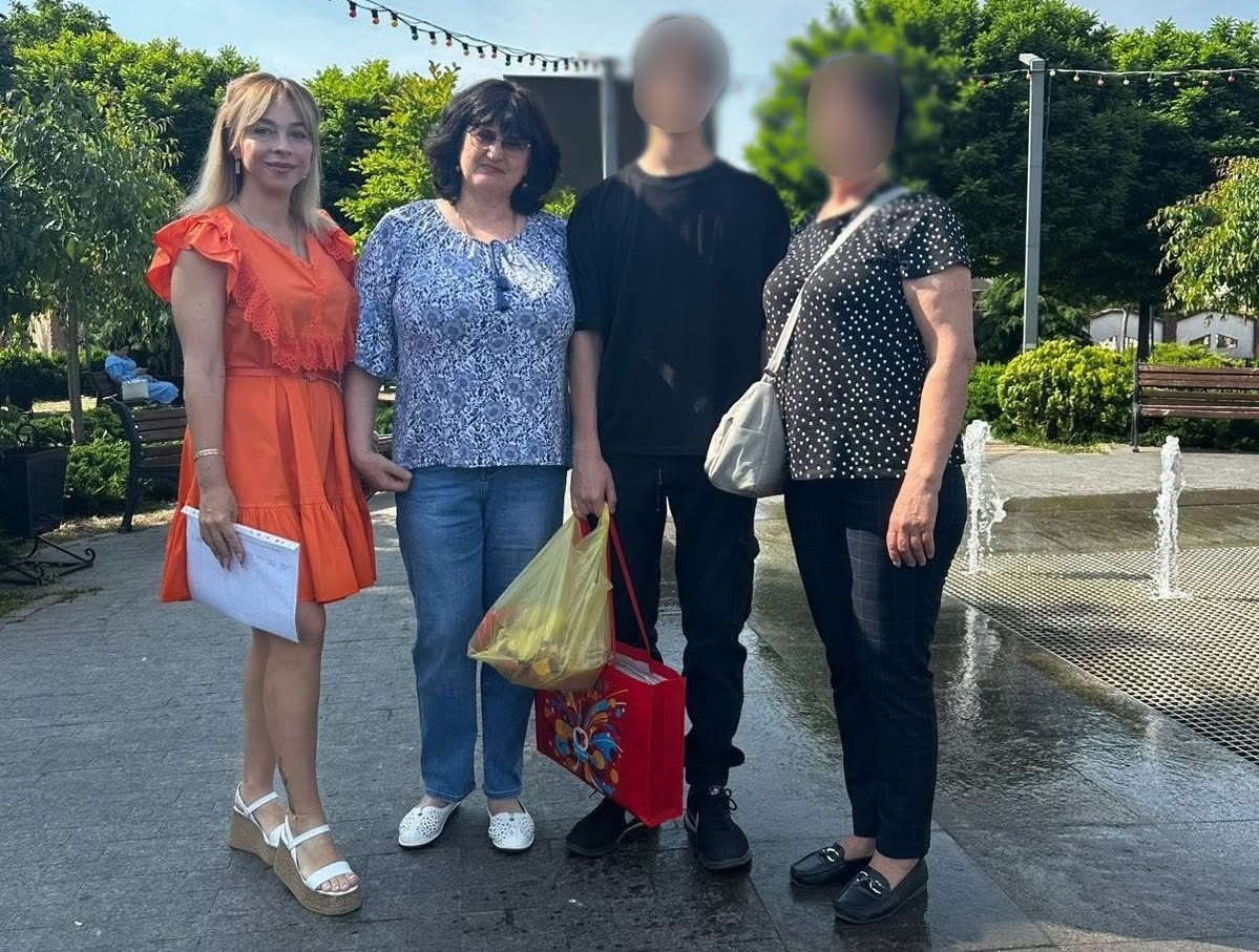 Чотири людини стоять в один ряд на брукованій доріжці в парку біля фонтана.  Ліворуч стоїть жінка в яскравій помаранчевій сукні та білих босоніжках на платформі, яка тримає в руках паперові документи.  Поруч із нею — жінка в блакитній блузці з візерунком та синіх джинсах.  По центру стоїть високий хлопець у чорній футболці та темних штанах, який тримає в руках великий жовтий пакет та червону подарункову сумку. Його обличчя розмите.  Праворуч стоїть жінка в чорній футболці в білий горошок та темних штанах, з розмитим обличчям та світлою сумкою через плече.  На задньому плані праворуч видно невисокі струмені води пішохідного фонтана, зелені дерева та паркову лавку.
