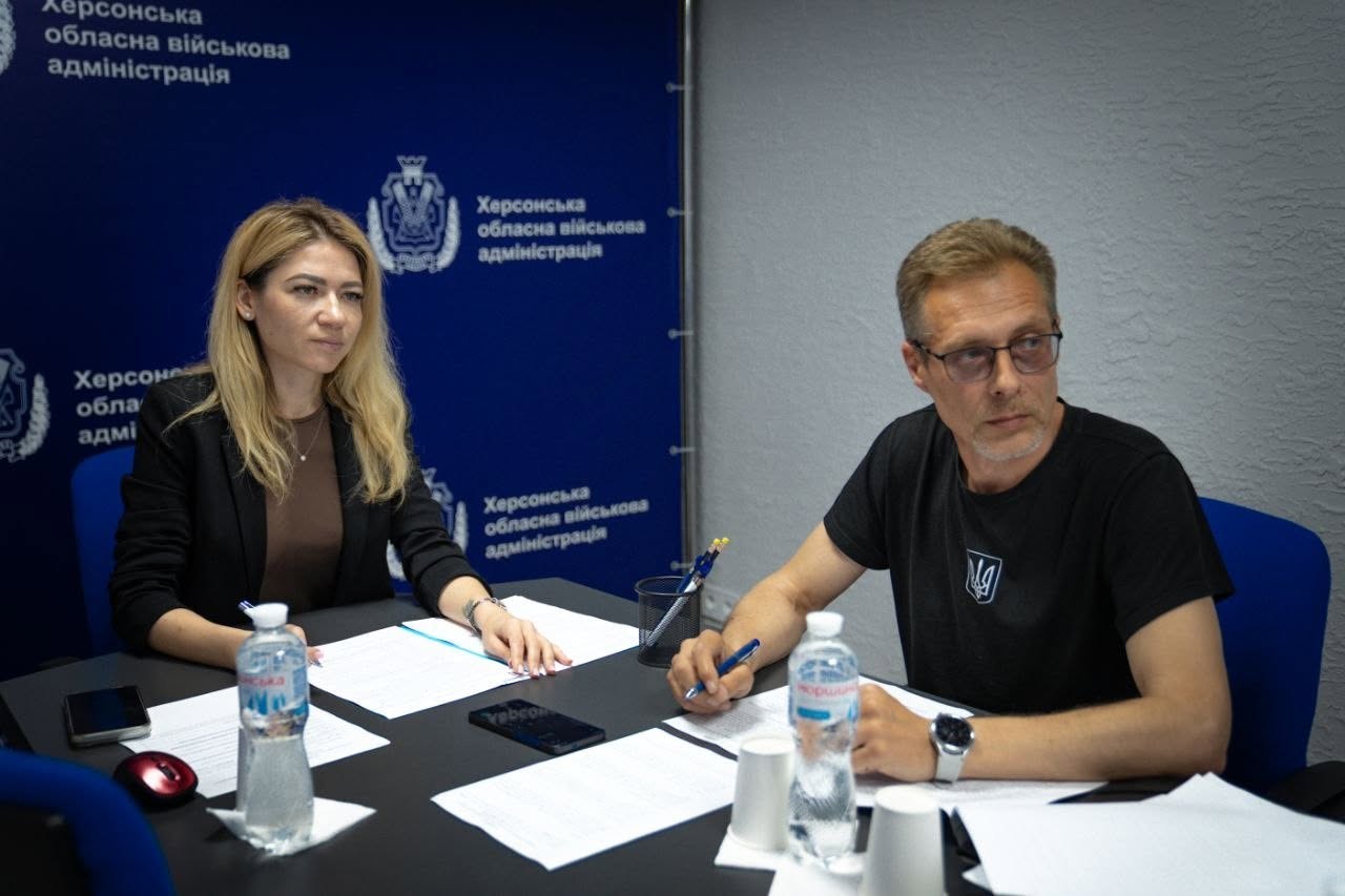 Двоє представників керівництва Херсонської ОВА беруть участь у засіданні за робочим столом. Ліворуч сидить Ольга Малярчук у чорному піджаку, перед якою розкладені папери. Праворуч за столом сидить чоловік в окулярах та чорній футболці з малим білим гербом України (Тризубом) на грудях. Він тримає в руці ручку, схилившись над роздрукованими документами.  На столі перед ними стоять дві пластикові пляшки води «Моршинська», паперові стаканчики, лежить мобільний телефон та підставка з канцелярією. Тло за спинами учасників розділене: ліва частина оформлена темно-синім банером «Херсонська обласна військова адміністрація», права — сірою рельєфною стіною кабінету.