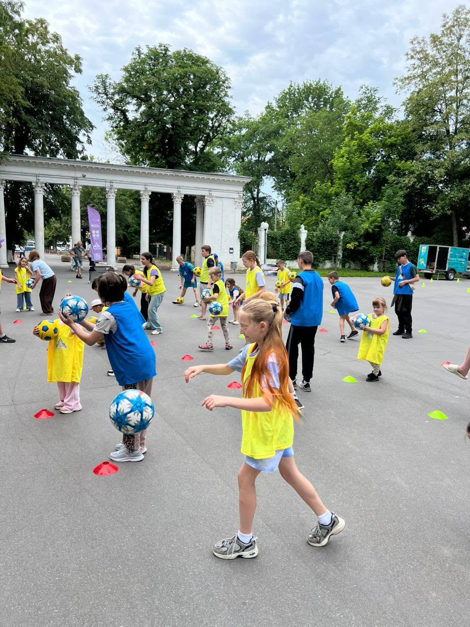 Динамічний кадр спортивної гри на відкритому повітрі. На передньому плані праворуч зображена довгокоса дівчинка у жовтій маніжці та блакитних шортах, яка біжить майданчиком. Лівіше від неї інша дитина у синій маніжці тримає в руках біло-блакитний футбольний м'яч, а біля її ніг у повітрі зафіксовано ще один такий самий м'яч. На задньому плані роззосереджені інші діти у жовтій та синій формі, які також тренуються з м'ячами. На асфальті розставлені різнокольорові пластикові фішки-конуси. Позаду видно білу класичну колонаду парку та зелені дерева.