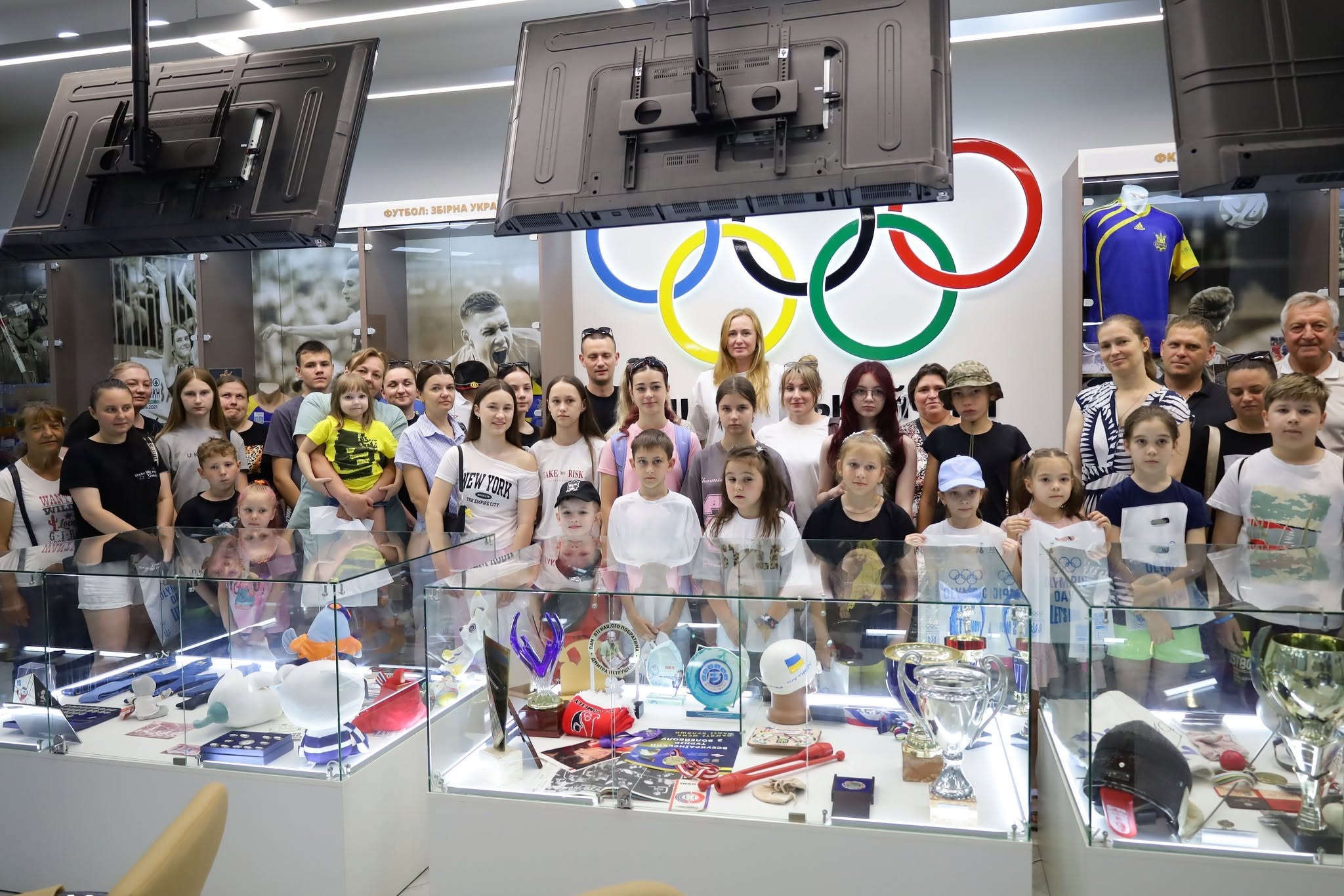 Велике групове фото всіх учасників екскурсії в інтер'єрі Олімпійського музею. Близько тридцяти людей — переважно діти, підлітки та кілька жінок — вишикувалися для знімка за трьома великими прямокутними скляними вітринами-тумбами. Всередині вітрин підсвічуються різноманітні кубки, кришталеві нагороди, спортивний інвентар (зокрема червоні булави для художньої гімнастики) та сувеніри. На білій стіні за спинами людей по центру закріплено великий кольоровий логотип у вигляді п'яти олімпійських кілець. Зверху над вітринами звисають три чорні монітори.