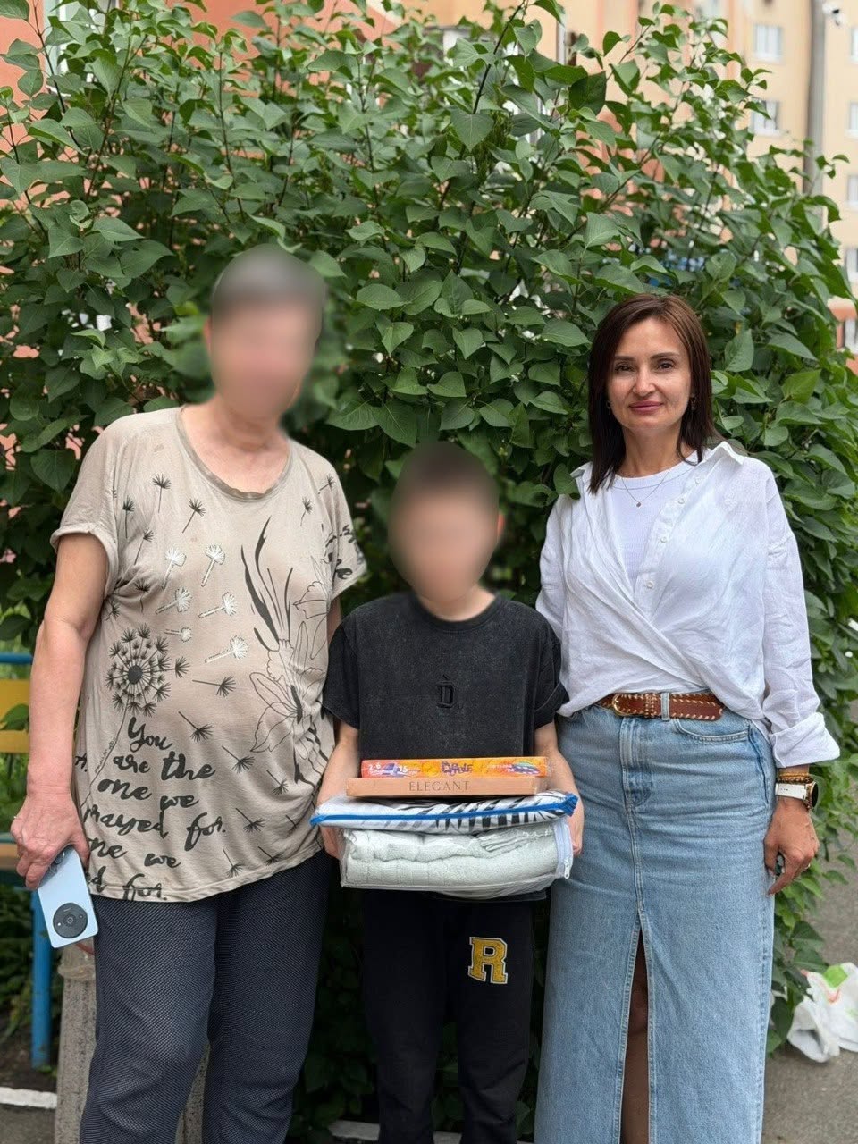 Три людини позують для фото на тлі великого зеленого куща біля житлового будинку.  Ліворуч стоїть жінка у світлій футболці з малюнком кульбаб і написами та темних штанах, яка тримає в руці мобільний телефон.  Праворуч стоїть жінка в білій блузці та довгій блакитній джинсовій спідниці з розрізом по центру та коричневим ременем.  Між ними по центру стоїть підліток у чорній футболці та чорних спортивних штанах з великою жовтою літерою «R». Він тримає в руках пакунок із новою постільною білизною чи ковдрою у прозорій упаковці, поверх якої лежать дві настільні ігри, зокрема коробка з написом «ELEGANT».  Обличчя жінки ліворуч та підлітка по центру повністю розмиті.