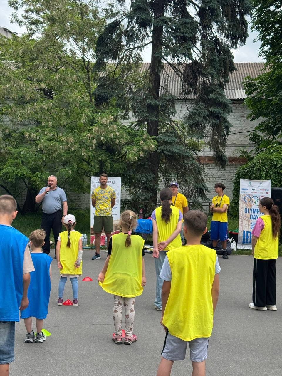 Офіційне відкриття або підбиття підсумків спортивного заходу. Група дітей у синіх та жовтих маніжках стоїть півколом на майданчику, повернувшись спиною до камери. Перед ними виступає старший чоловік у сірій футболці-поло, який говорить у мікрофон. Поруч із ним стоїть молодий тренер у патріотичній жовто-блакитній футболці. Позаду них ростуть високі хвойні та листяні дерева, а праворуч встановлено вертикальний білий банер із символікою Олімпійських ігор та великим написом «OLYMPIC DAY. LET'S MOVE».