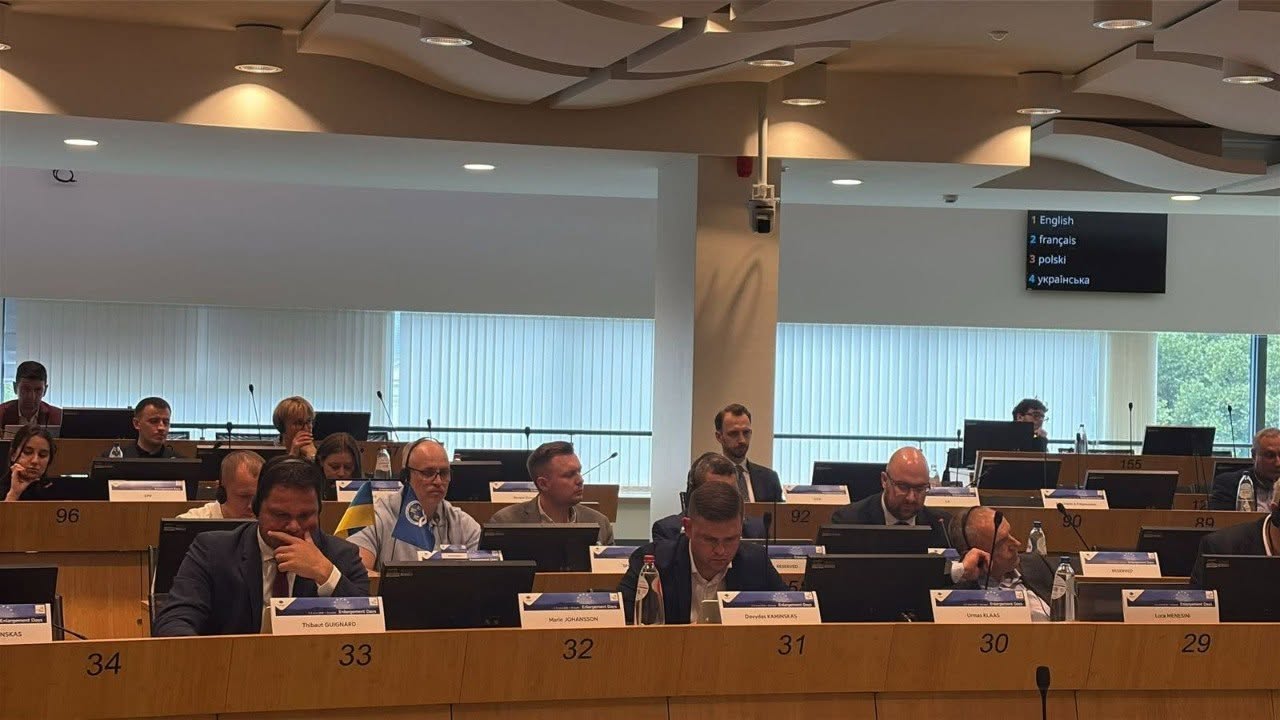 Панорамний знімок великої зали засідань під час офіційної європейської конференції. Кілька рядів делегатів у ділових костюмах сидять за довгими напівкруглими дерев'яними столами, обладнаними моніторами, мікрофонами та іменними табличками. На столах перед учасниками встановлені номери місць: «34», «33», «32», «31», «30», «29». Деякі з присутніх роблять нотатки або слухають виступи через навушники. На задньому плані по центру видно велике вікно з вертикальними жалюзі, а праворуч угорі під стелею закріплено чорне електронне табло з переліком мов перекладу, де четвертим пунктом вказано «українська».