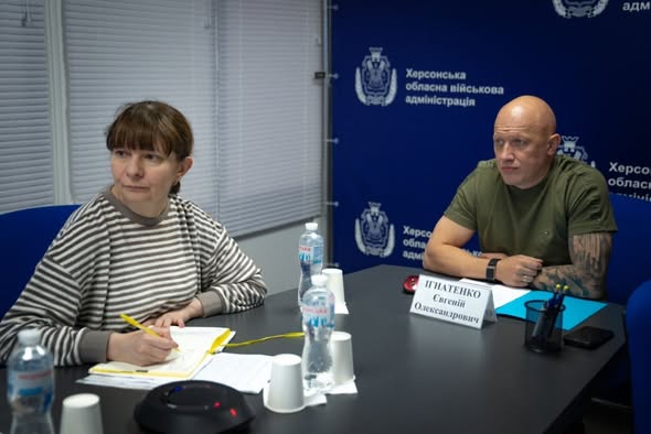 Робоча зустріч у кабінеті під час онлайн-зв'язку. За темно-сірим столом сидять двоє представників Херсонщини. Ліворуч — жінка у смугастій кофті, яка робить нотатки жовтою ручкою у блокноті. Праворуч — лисий чоловік у військовій футболці кольору хакі з татуюванням на лівій руці. Перед ним на столі встановлено білу табличку з написом «ІГНАТЕНКО Євгеній Олександрович», лежить папка із блакитними паперами, мобільний телефон та підставка з ручками. Також на столі розставлені пластикові пляшки з водою «Моршинська» та білі паперові стаканчики. На задньому плані праворуч розташовано суцільний темно-синій банер із повторюваними білими логотипами та написом «Херсонська обласна військова адміністрація».