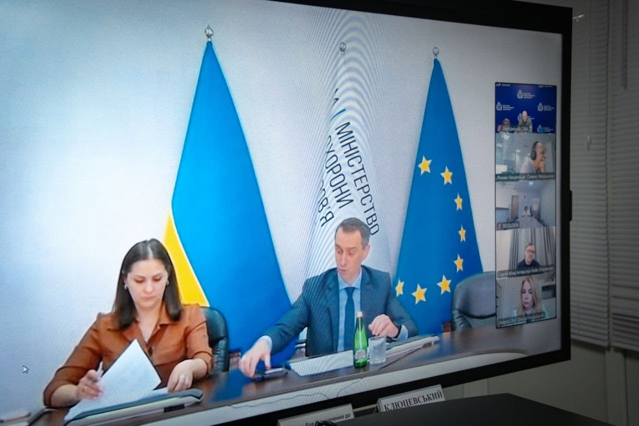 Світлина великого плаского монітора, на якому транслюється селекторна онлайн-нарада. У центрі екрана у великому головному вікні засідають двоє представників відомства. Чоловік у синьому костюмі та краватці сидить за столом, на якому стоїть пляшка води та склянка; позаду нього встановлено державний прапор України, білий прапор із написом «МІНІСТЕРСТВО ОХОРОНИ ЗДОРОВ'Я» та прапор Європейського Союзу. Ліворуч від нього за столом сидить жінка в коричневій блузці, яка переглядає паперові документи. У правій частині монітора вертикальною стрічкою виведені п'ять менших вікон із трансляціями інших учасників відеоконференції. У самому низу екрана видно табличку з прізвищем «КЛЮЦЕВСЬКИЙ».