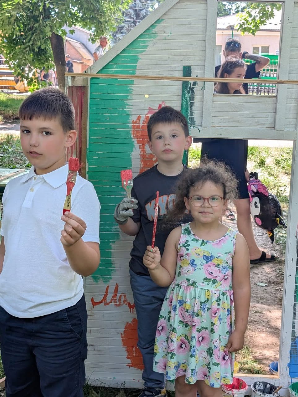 Три дитини позують перед камерою на майданчику, тримаючи в руках пензлики для малювання з червоною фарбою. Ліворуч стоїть хлопчик у білій футболці-поло та темних штанях, який тримає пензлик вертикально. По центру стоїть другий хлопчик у чорній футболці з малюнком та робочій рукавиці на правій руці. Праворуч на передньому плані посміхається дівчинка в окулярах та світлій сукні в рожево-жовтий квітковий принт. Позаду дітей розташована дерев'яна ігрова конструкція, частково пофарбована зеленою та помаранчевою фарбою, у вікні якої видно розмиті силуети дорослих.