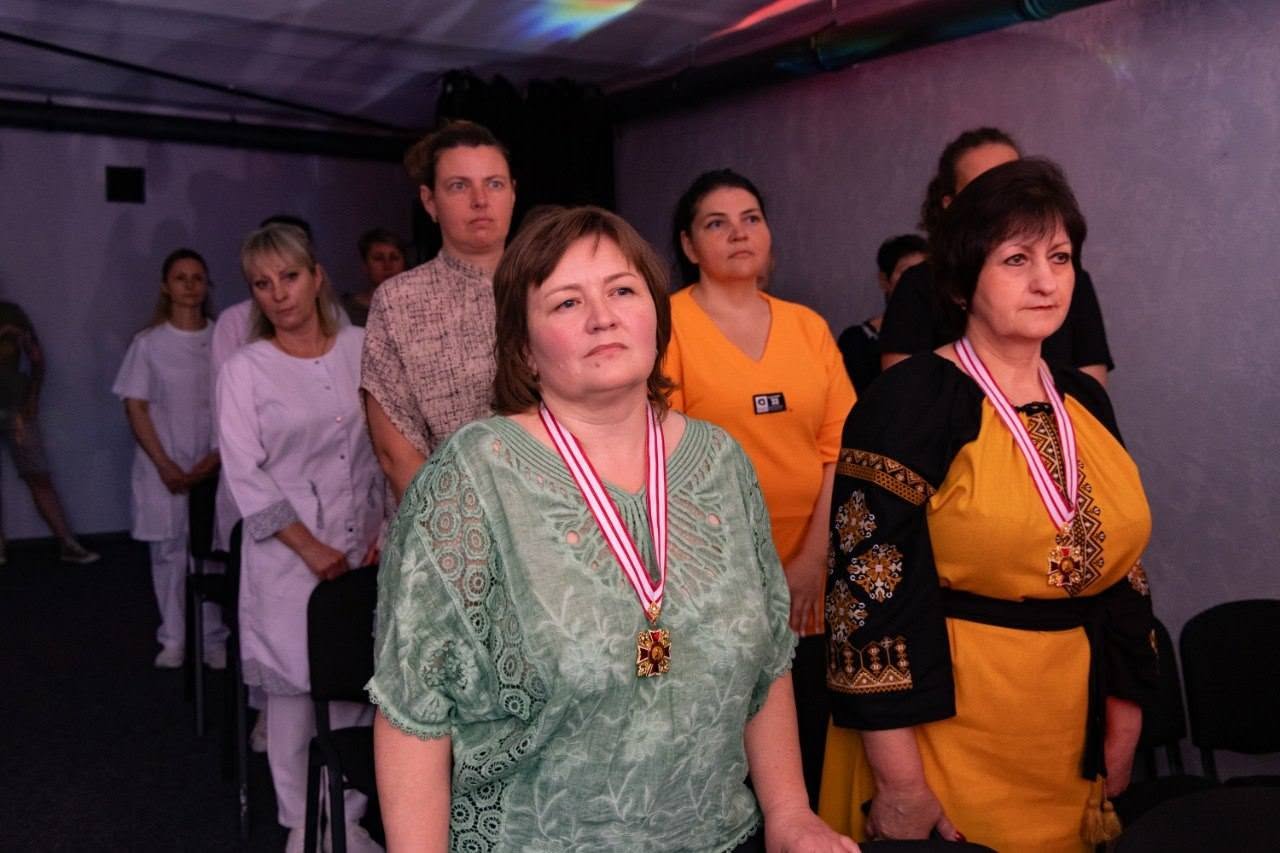 Ряд медичних працівників та відвідувачів стоїть у залі під час урочистої частини заходу. На передньому плані праворуч стоїть жінка у жовто-чорній сукні з традиційним вишитим орнаментом на рукавах, на її шиї на червоно-білій стрічці висить металевий орден. Поруч із нею ліворуч стоїть інша жінка у світло-зеленій блузці з аналогічним орденом на шиї.  У наступних рядах видно жінку в помаранчевій футболці, жінку в бежевій блузці, а також медичних працівників у білих медичних халатах і костюмах. Обличчя присутніх повернуті вправо та вгору в бік сцени. Зала має приглушене фіолетове освітлення.