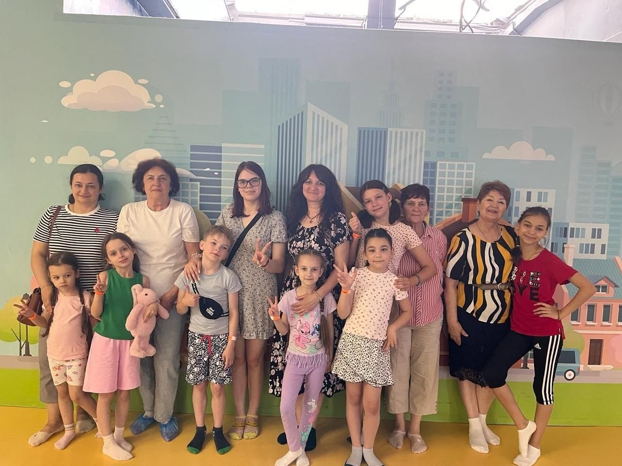 Групове фото одинадцяти учасниць заходу — п'яти жінок та шести дівчаток і хлопчиків різного віку. Вони вишикувалися в один ряд і позують для знімка, дехто з дітей показує пальцями жест «вікторія» (V) або тримає руки на поясі. Вони одягнені в літній повсякденний одяг: футболки, шорти, легкі сукні та спідниці. У руках однієї з дівчаток ліворуч — рожева плюшева іграшка-зайчик. Група стоїть на тлі яскравої фотозони — стіни з пастельним малюнком сучасного міста з висотними будинками, хмарами та повітряною кулею в небі. Підлога в цій зоні має суцільне насичено-жовте покриття.