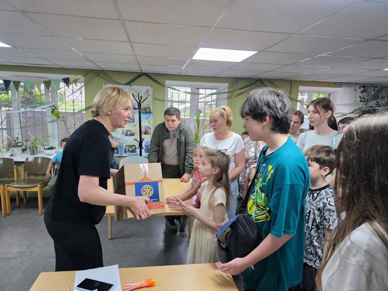 Момент видачі гуманітарної допомоги чи подарунків у приміщенні центру. Ліворуч світловолоса жінка в чорній футболці передає велику картонну коробку маленькій дівчинці в бежевій сукні. На коробці наклеєно круглу емблему міжнародної організації «Rotary International» із зображенням пожежної машини. Навколо них стоять та спостерігають інші діти й дорослі, зокрема хлопець у бірюзовій футболці з чорною сумкою через плече та чоловік у куртці захисного кольору. На передньому плані видно куточок дерев'яного столу з мобільним телефоном та кольоровими олівцями.