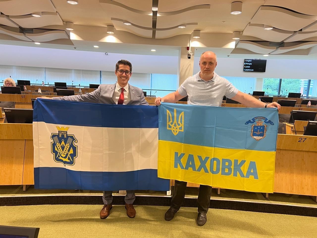 Два чоловіки стоять у повний зріст по центру зали засідань і спільно розгортають два великі офіційні прапори. Ліворуч чоловік у сірому костюмі, білій сорочці та червоній краватці тримає синьо-білий прапор із гербом Херсонської області по центру. Праворуч чоловік у світло-блакитному поло та темних штанях тримає державний прапор України, на нижній жовтій половині якого нанесено великий синій напис «КАХОВКА», а у верхньому правому кутку розміщено герб міста Каховка. Обидва учасники посміхаються і дивляться в об'єктив камери на тлі амфітеатру порожніх робочих місць делегатів.
