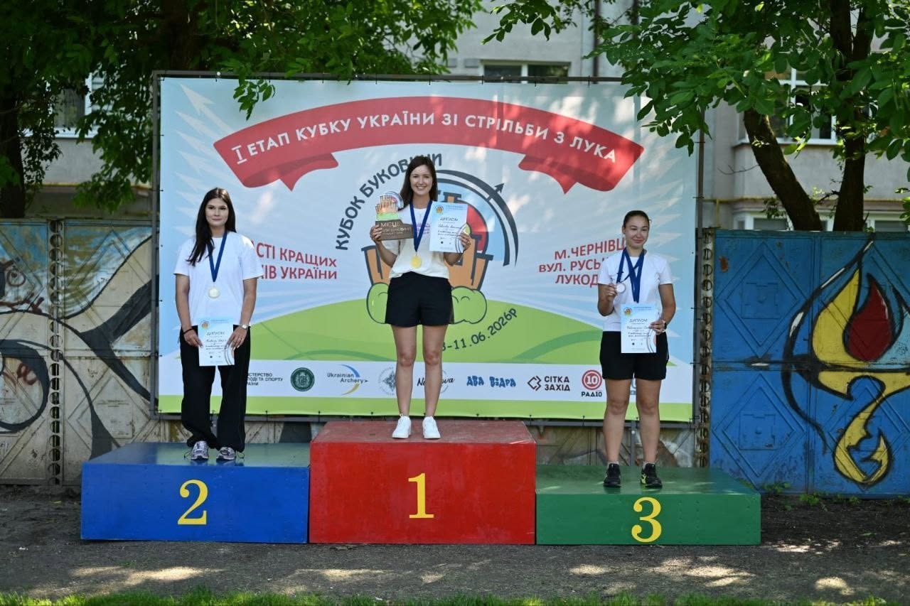 Три переможниці змагань зі стрільби з лука стоять на трирівневому кольоровому п'єдесталі пошани на тлі великого інформаційного банера під відкритим небом. По центру на найвищій червоній сходинці з цифрою «1» стоїть дівчина у білій футболці та чорних шортах, тримаючи в руках кубок та диплом. Ліворуч на синій сходинці з цифрою «2» стоїть дівчина у білій футболці та чорних спортивних штанях із дипломом у руці та срібною медаллю на шиї. Праворуч на зеленій сходинці з цифрою «3» стоїть третя призерка у білій футболці та чорних шортах із дипломом та бронзовою медаллю. На банері позаду них, окрім офіційних назв етапу Кубку України та Кубку Буковини в Чернівцях, нанесено дати проведення: «08-11.06.2026р.». З обох боків від банера видно зелені дерева, ліворуч — бетонний паркан із графіті, праворуч — синя металева брама.