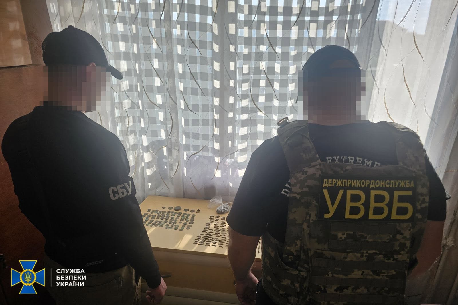 Двоє правоохоронців зафіксовані зі спини у кімнаті перед столом із вилученими речовими доказами.  Ліворуч стоїть співробітник у чорній кофті з великим білим написом «СБУ» на лівому плечі та в чорній кепці.  Праворуч стоїть представник у камуфляжному бронежилеті-розвантаженні, на спині якого закріплено велику нашивку з жовтим текстом: «ДЕРЖПРИКОРДОНСЛУЖБА УВВБ» (Управління внутрішньої та власної безпеки).  Обидва чоловіки дивляться вниз на розкладені перед ними на столі старовинні монети та наконечники стріл. Обличчя та голови правоохоронців повністю розмиті цифровим піксельним фільтром. На задньому плані видно вікно з білою квадратною та візерунчастою тюлю.