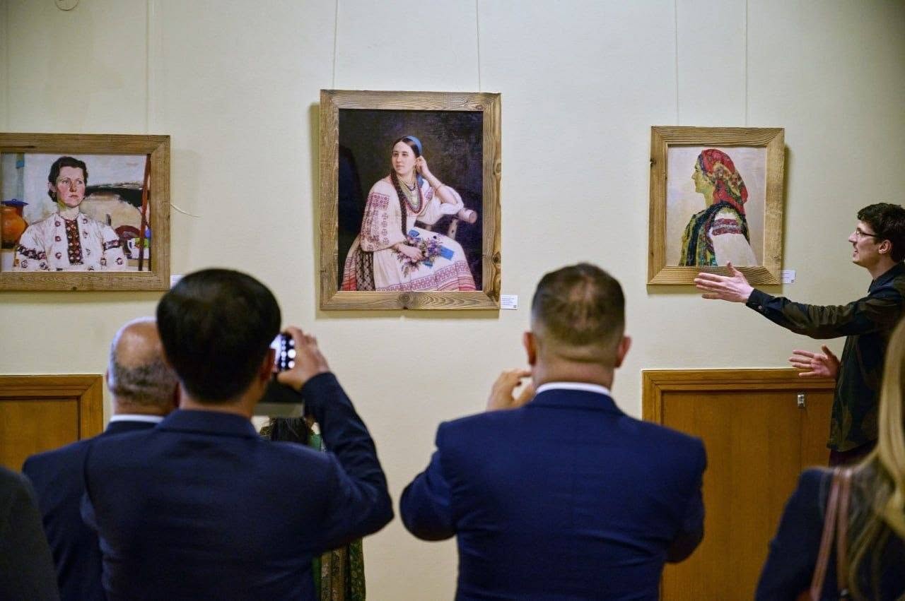 Дипломати та учасники міжнародної делегації слухають екскурсовода, стоячи у виставковому залі. У центрі кадру стоїть жінка в зеленому традиційному костюмі з орнаментом, склавши руки перед собою. Ліворуч від неї — чоловік у чорному костюмі та білій сорочці, а також дівчина у сукні в дрібний чорно-білий горошок. Праворуч стоїть дівчина у білій блузці та світлих штанях, тримаючи сонцезахисні окуляри на вирізі сорочки. Позаду них стоять інші члени делегації — чоловік у синьому піджаку, жінка в темному жакеті та чоловіки в костюмах з краватками. На стіні праворуч висить картина у дерев'яній рамі. Підлога в приміщенні вистелена геометричним дерев'яним паркетом.