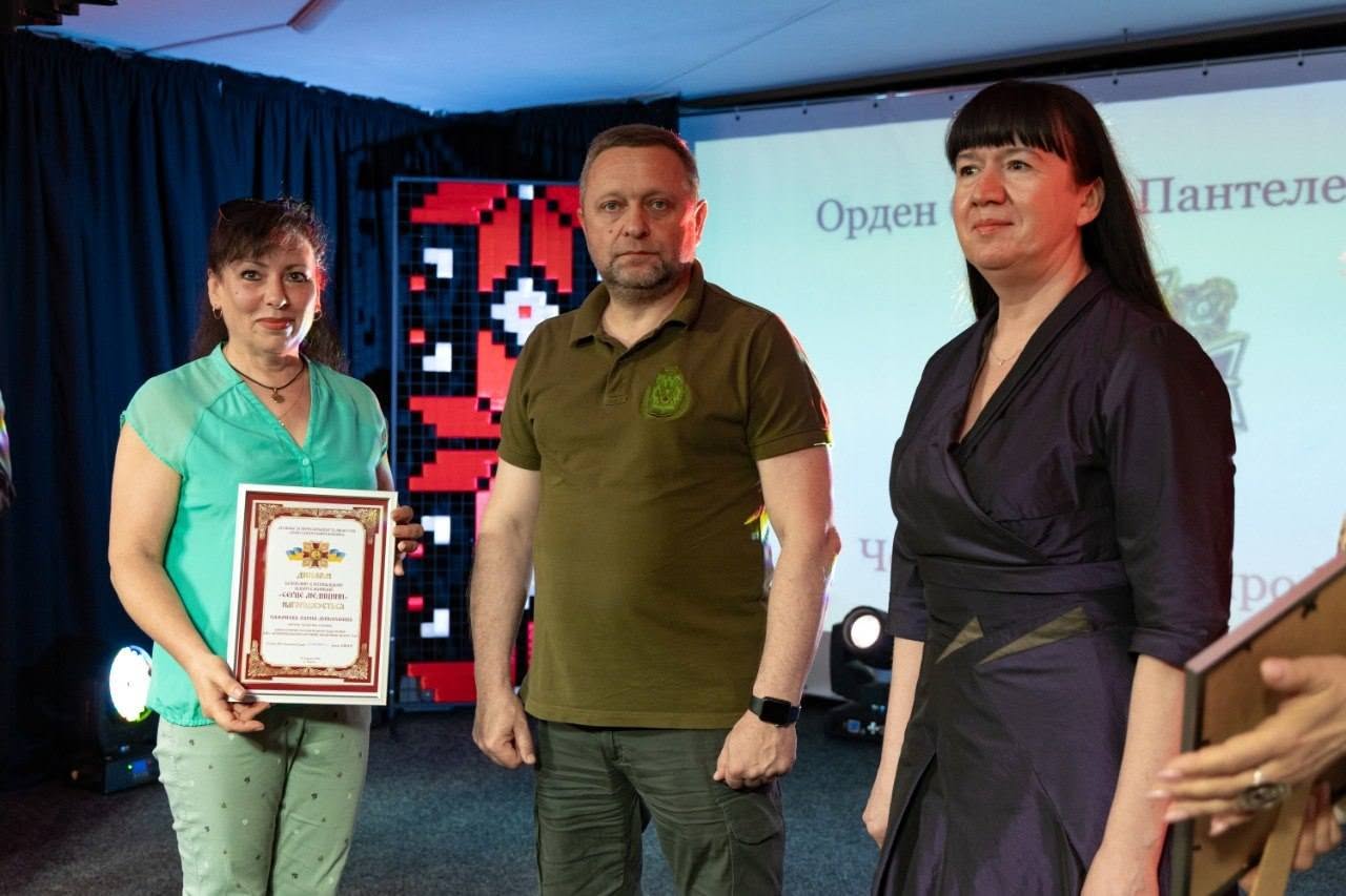 Момент вручення диплома на сцені. Ліворуч у напівпрофіль стоїть жінка-лауреат у світло-бірюзовій блузці та світлих штанях із дрібним візерунком, яка тримає в руках нагородний диплом у рамці.  По центру стоїть заступник начальника ОВА Дмитро Бутрій у мілітарі-поло, спрямувавши погляд перед собою. Праворуч від нього у профіль зафіксована жінка в темно-синій сукні. У правому нижньому кутку кадру видно розмиті руки іншої людини, яка передає або тримає ще одну нагороду в дерев'яній рамці. На задньому плані світиться екран із написом «Орден ... Пантеле...».