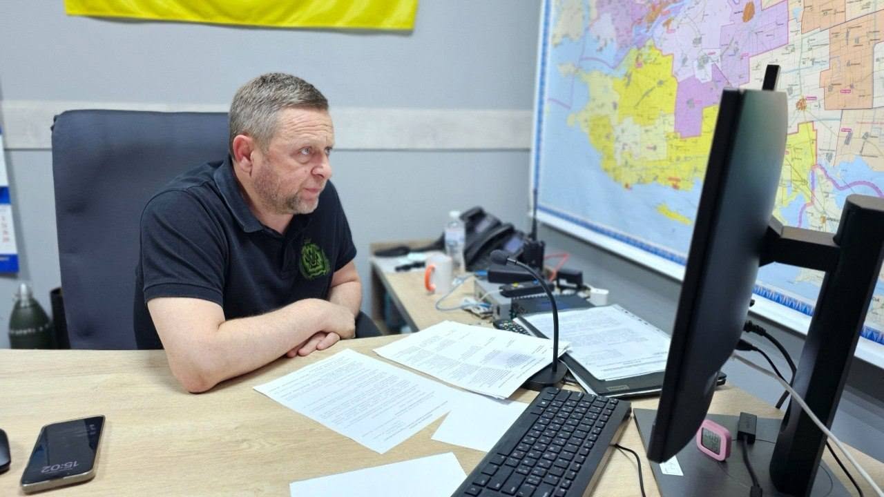 Перший заступник начальника Херсонської ОВА Дмитро Бутрій бере участь у робочій онлайн-нараді у своєму службовому кабінеті. Чоловік сидить у профілі ліворуч у чорному офісному кріслі, він одягнений у чорну футболку-поло з зеленим круглим гербом на грудях та зосереджено дивиться на великий чорний монітор комп'ютера.  На світлому дерев'яному столі перед ним розкладено паперові документи, стоїть комп'ютерна миша, чорна клавіатура, лежить смартфон та встановлено настільний мікрофон на гнучкій ніжці. На задньому плані праворуч на стіні закріплено велику кольорову карту Херсонської області зі схематичним поділом на громади. У лівому верхньому кутку видно частину жовтого полотна державного прапора України, а на столі на другому плані — робочі папери, канцелярію та стаціонарний телефон.