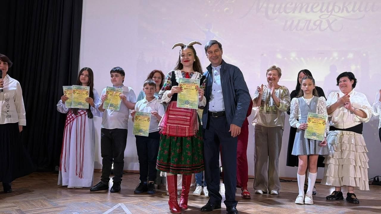 Учасники та організатори фестивалю вишикувалися в ряд на сцені для пам'ятного фото.  По центру стоїть дівчина в яскравому українському національному костюмі з вишивкою, червоним фартухом, зеленою спідницею та оригінальним головним убором з імітацією ріжок. Вона тримає диплом фестивалю.  Праворуч від неї стоїть чоловік у синій куртці та білій сорочці, який посміхається.  З обох боків від них стоять інші лауреати — діти та дорослі жінки в народних строях, сукнях та вишиванках, дехто з них також тримає нагородні дипломи, а інші аплодують.  На білому екрані ззаду розмито відображається назва заходу «Мистецький шлях». Підлога сцени має дерев'яну паркетну текстуру.