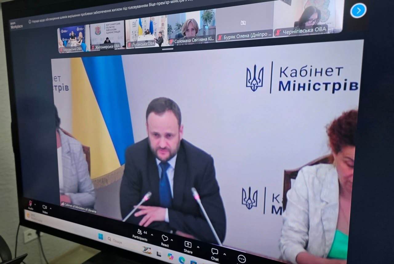 Екран великого комп'ютерного монітора під кутом, на якому відображається перебіг онлайн-наради у програмі Zoom. Головне центральне вікно займає трансляція з Кабінету Міністрів України, де за столом на тлі державного прапора та логотипу «Кабінет Міністрів» сидітиме чоловік у темному костюмі, сорочці та краватці, який виступає перед мікрофоном. З обох боків від нього видно інших учасників урядової президії.  У верхній горизонтальній стрічці монітора виведені вікна інших підключених учасників конференції:  «Херсонська ОВА» (видно представників адміністрації у кабінеті);  «Житомирська ОВА»;  «Луганська ОВА»;  «Соломаха Світлана Кі...»;  «Буряк Олена (Дніпро...»;  «Чернігівська ОВА».  Нижня панель програми містить стандартні інструменти керування конференцією: кількість учасників (44), кнопки «React», «Share», «Chat».