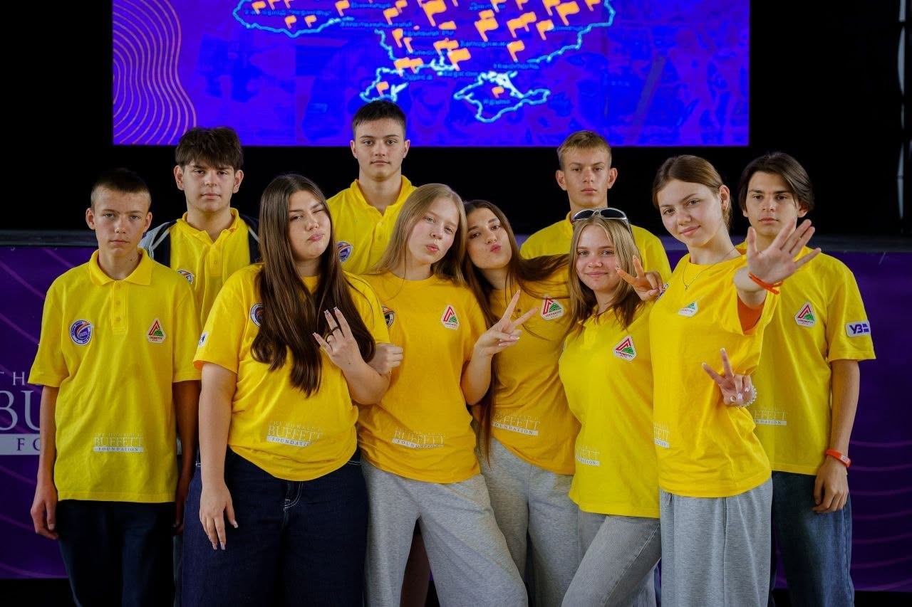 Спільне групове фото дев'яти підлітків (чотирьох хлопців та п'яти дівчат) на сцені у приміщенні. Вони вишикувані у два ряди: хлопці стоять позаду, дівчата — попереду. Усі підлітки одягнені в уніфіковані жовті футболки та сірі або сині повсякденні штани й джинси.  Учасники заходу весело позують: показують пальцями жести «вікторія», махають руками у кадр та посміхаються. Позаду них на великому мультимедійному екрані горить яскраве синє тло з контурною картою України, на якій жовтими прапорцями позначено різні локації чи міста.