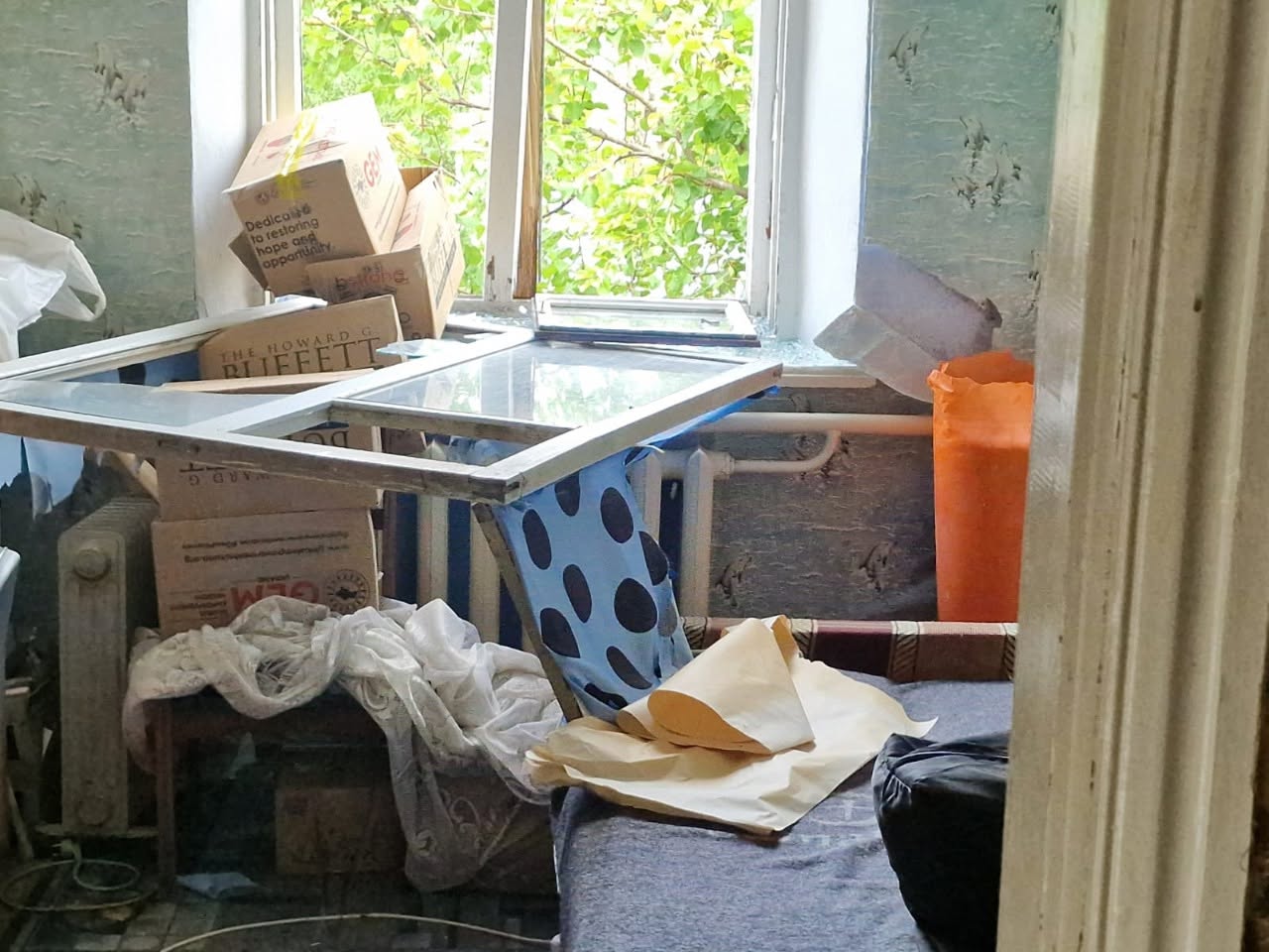 Робоча група спілкується з постраждалими мешканцями на вулиці біля пошкодженого під час обстрілу подвір'я. У центрі кадру чоловік у сірій футболці та джинсах розмовляє з Ольгою Малярчук, яка стоїть спиною до камери в білій кофті. Поруч знаходяться ще дві жінки та чоловік у синій сорочці. На задньому плані росте велике зелене дерево, за яким видно одноповерховий будинок із понівеченим дахом, паркан та ворота. Ліворуч на дорозі припарковано білий мікроавтобус екстрених служб.