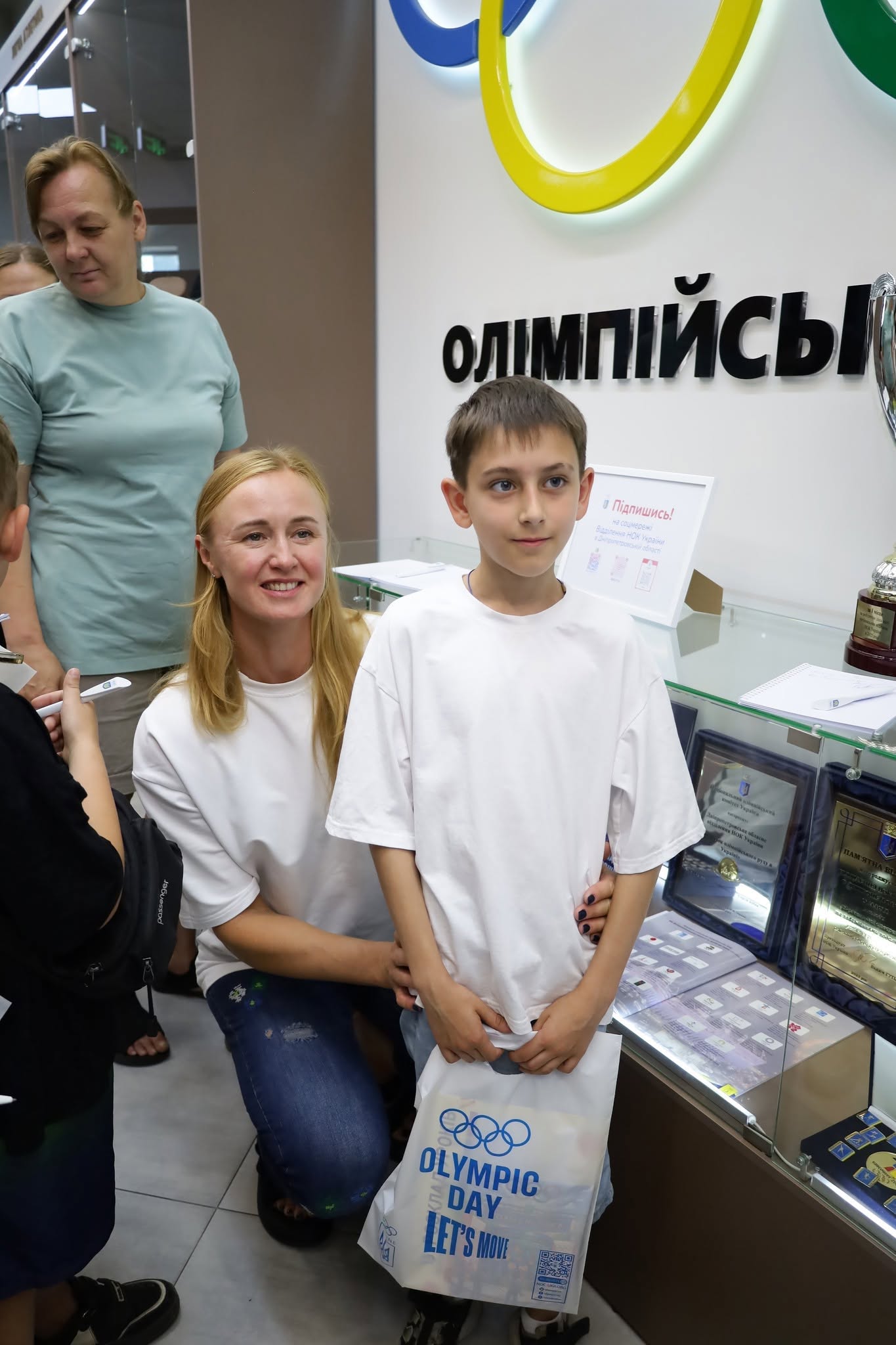 Світловолоса жінка у білій футболці та синіх джинсах присіла поруч із хлопчиком років десяти, який одягнений у вільну білу футболку та тримає в руках білий пакет із синім логотипом «OLYMPIC DAY. LET'S MOVE». Вони позують перед скляною вітриною музею, всередині якої виставлені нагороди, медалі та пам'ятні плакетки. На задньому плані на білій стіні закріплено об'ємний чорний напис «ОЛІМПІЙСЬК...» під великим жовтим олімпійським кільцем. На вітрині також стоїть інформаційна картка з червоним написом «Підпишись!».