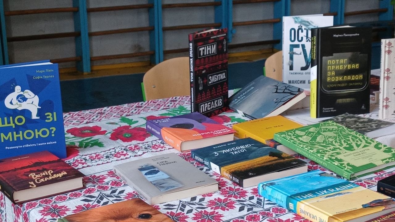 Книжкова розкладка на столі, застеленому білою скатертиною з традиційним українським червоно-чорним орнаментом. На передньому та середньому планах розкладено понад десять сучасних художніх та документальних книг у різнокольорових обкладинках. Серед них чітко видно видання: «Що зі мною?», «Вечір у Стамбулі», «З любов'ю — тато!», «Потяг прибуває за розкладом», «Тіні забутих предків» та книга «THE UKRAINE». На задньому плані видно дерев'яну шведську стінку блакитного кольору та стільці.