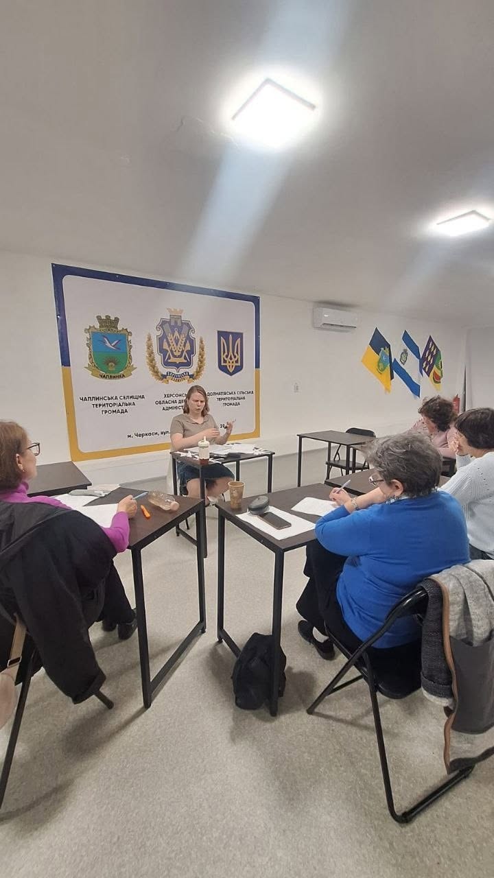 Групове інформаційне заняття для жінок у світлому кабінеті центру допомоги. За окремими невеликими столами з чорними металевими ніжками сидять відвідувачки. На передньому плані праворуч боком до камери сидить жінка у яскраво-синьому светрі, яка робить записи в зошиті. Навпроти присутніх за окремим столом сидить молода жінка-лектор у бежевій футболці, яка емоційно пояснює матеріал, жестикулюючи руками.  На білій стіні за її спиною закріплено великий офіційний банер із зображенням гербів та написами: «ЧАПЛИНСЬКА СЕЛИЩНА ТЕРИТОРІАЛЬНА ГРОМАДА», «ХЕРСОНСЬКА ОБЛАСНА ДЕРЖАВНА АДМІНІСТРАЦІЯ» та «ДОЛМАТІВСЬКА СІЛЬСЬКА ТЕРИТОРІАЛЬНА ГРОМАДА. м. Черкаси, вул. Остафія Дашковича, 39». Праворуч на стіні висять три прапори.