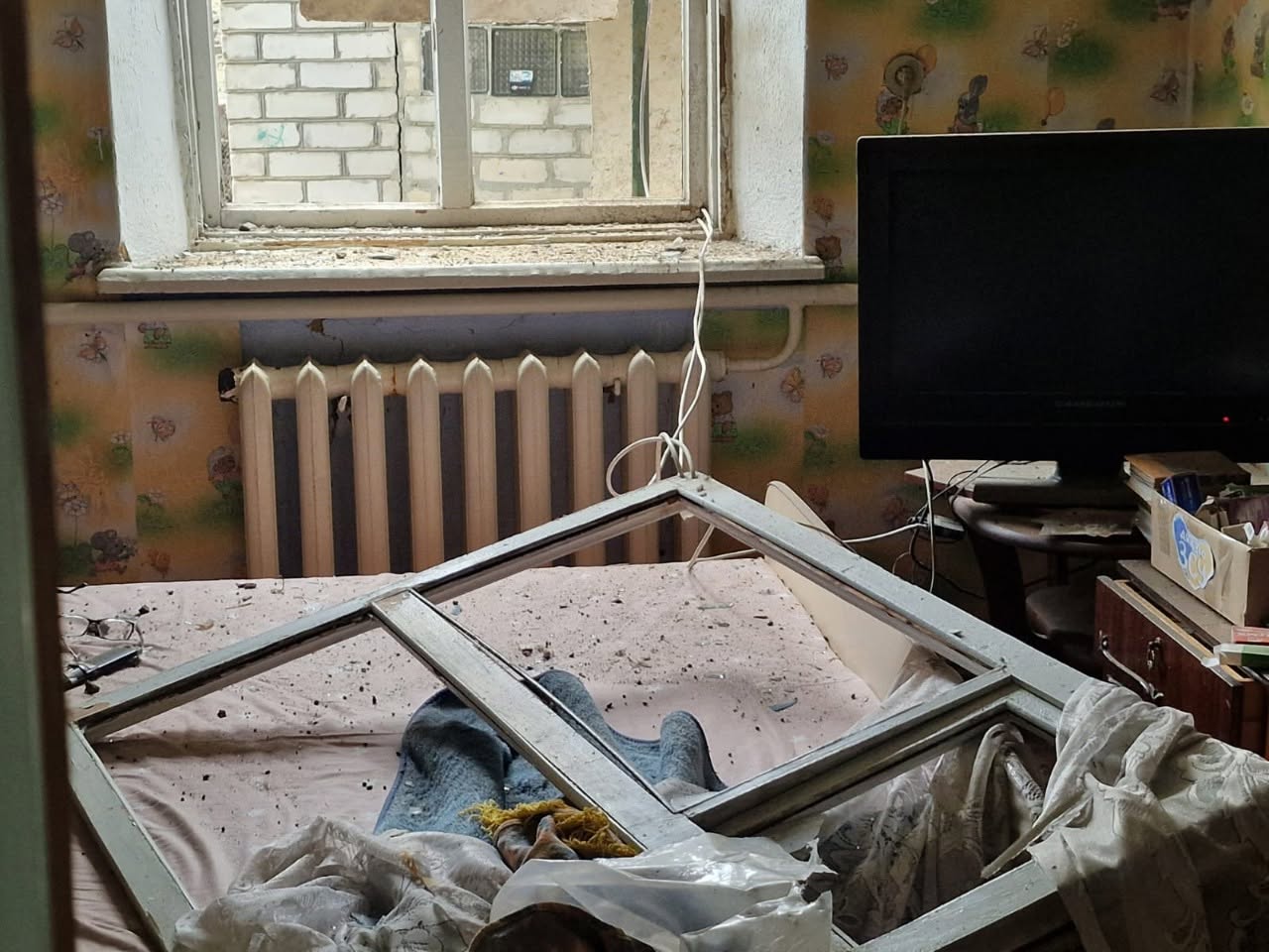 Заступниця начальника Херсонської ОВА Ольга Малярчук разом із місцевим літнім мешканцем оглядає пошкоджений приватний будинок з білої силікатної цегли. Постраждалий чоловік у сірій футболці з написом «PERFECT» тримається рукою за поручні металевих сходів і щось розповідає. Ольга Малярчук, одягнена у білу спортивну кофту з капюшоном, зосереджено слухає його. На задньому плані видно вибите вікно будинку, завішене пошкодженою тюлю, та частково зруйнований дах.