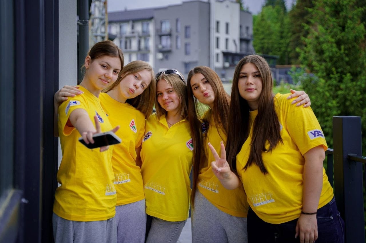 П'ять дівчат-підлітків позують разом для фотографії на відкритій терасі. Усі вони одягнені в однакові яскраві жовті футболки-поло з круглими нашивками на грудях та принтом «The Howard G. Buffett Foundation» на нижній частині. Дівчата стоять впритул одна до одної, посміхаються та обіймаються за плечі.  Дівчина ліворуч витягнула руку вперед, імітуючи зйомку селфі на смартфон, та показує пальцями жест «вікторія» (V). Інша дівчина, яка стоїть праворуч, також підняла руку з цим жестом. На розмитому задньому плані видно сучасні багатоповерхові житлові будинки сірого кольору та зелені дерева.