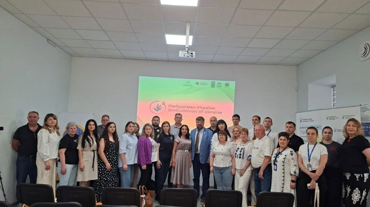 Велика група людей (близько тридцяти учасників заходу) вишикувалася для спільного фото в один ряд у світлому конференц-залі. Серед присутніх — чоловіки та жінки різного віку у діловому, вишиваному та повсякденному літньому одязі. На передньому плані перед людьми видно спинки порожніх чорних офісних крісел.  На задньому плані по центру світиться великий мультимедійний екран із градієнтним червоно-зеленим тлом, логотипом у вигляді долоні з птахом та офіційним двомовним написом: «Омбудсман України / Ombudsman of Ukraine». Праворуч біля стіни встановлено білі інформаційні ролапи (стенди) з логотипами відомства. Зала обладнана підвісною стелею типу «Армстронг» із квадратними лампами.