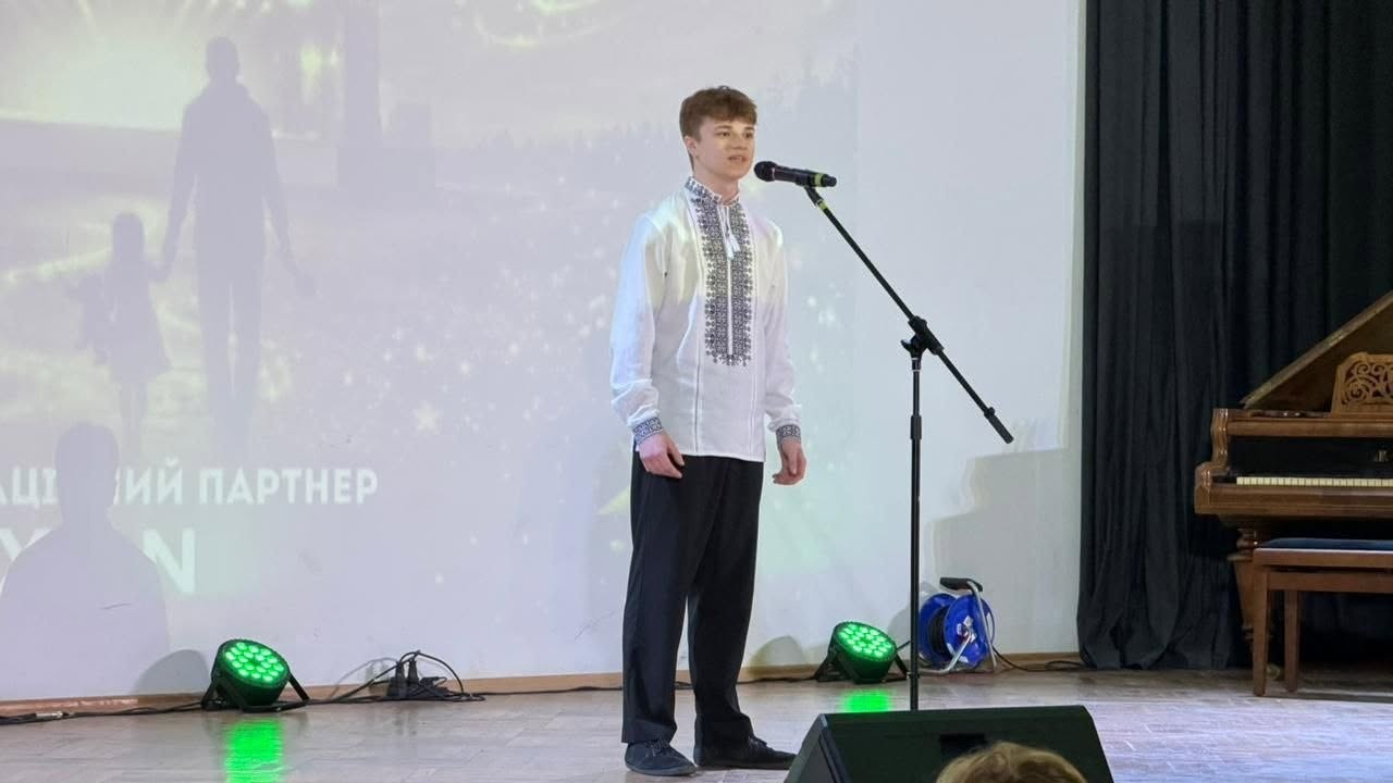 Молодий хлопець-виконавець виступає на сцені залу. Він одягнений у традиційну білу українську вишиванку з сіро-чорним орнаментом на грудях та манжетах і темні штані. Юнак стоїть перед стійкою з мікрофоном, спрямувавши погляд у зал під час виступу.  За його спиною на великому білому екрані відображається світла проєкція із силуетами дорослого й дитини, які йдуть за руку, та написами «...ій партнер». На підлозі сцени встановлено два невеликі круглі прожектори, які підсвічують простір зеленим світлом. Праворуч на задньому плані видно частину коричневого дерев'яного рояля.