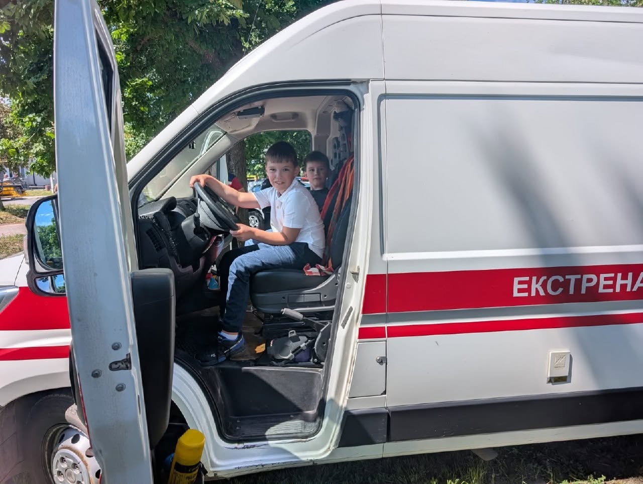Два хлопчики сидять усередині кабіни білого автомобіля екстреної медичної допомоги через відкриті водійські двері. Хлопчик на передньому плані у білій футболці та синіх джинсах тримає руки на чорному кермі автомобіля, імітуючи керування, і посміхається в об'єктив. Позаду нього на пасажирському сидінні видно другого хлопчика. На білому борті спецтранспорту нанесено широку червону горизонтальну смугу з великим білим написом «ЕКСТРЕНА». Машина стоїть на вулиці біля газону, на передньому плані у дверях видно жовтий балончик.
