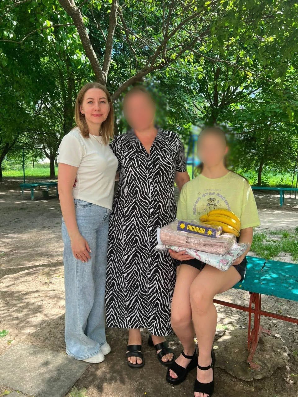 Три людини зафіксовані у повний зріст на тлі густих зелених дерев у парку чи на дитячому майданчику.  Ліворуч стоїть жінка у білій футболці та світло-блакитних широких джинсах.  Поруч із нею по центру стоїть жінка в довгій чорно-білій сукні на ґудзиках із косим анімалістичним принтом та чорних босоніжках.  Праворуч на синій дерев'яній лавці сидить дівчина-підліток у світло-жовтій футболці з малюнком та чорних шортах. Вона тримає на колінах пакунок із речами в прозорій плівці, коробку цукерок «PICHKAR» та велику зв'язку стиглих жовтих бананів.  Обличчя жінки по центру та дівчини на лавці повністю розмиті. На задньому плані видно металеві конструкції дитячого майданчика.