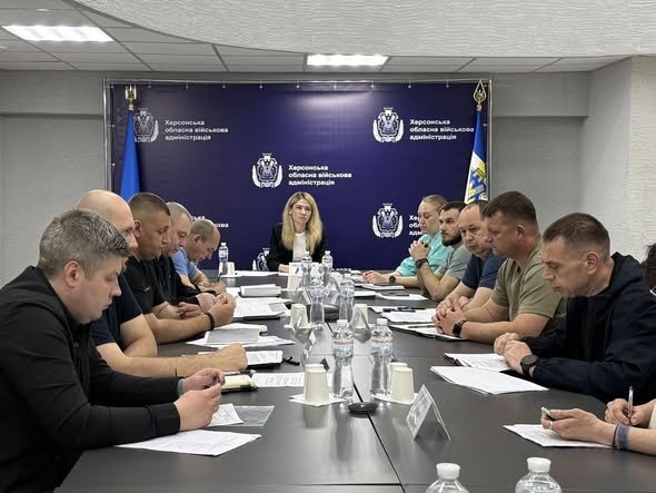 Робоча нарада керівництва та представників підрозділів у конференц-залі Херсонської ОВА. Близько п'ятнадцяти учасників засідають за довгим Т-подібним столом темно-сірого кольору, що заставлений документами, пляшками з водою та паперовими стаканчиками. По центру столу розміщено мікрофони "жабки".  У торці столу на чолі засідання сидить заступниця начальника ОВА Ольга Малярчук. Чоловіки та жінки вздовж столу вивчають документи або роблять нотатки. За спиною головуючої встановлено великий темно-синій пресволл із написом «Херсонська обласна військова адміністрація», прикрашений гербами області, прапором України та синім прапором відомства. Зала має світлі стіни та вбудоване стельове освітлення.