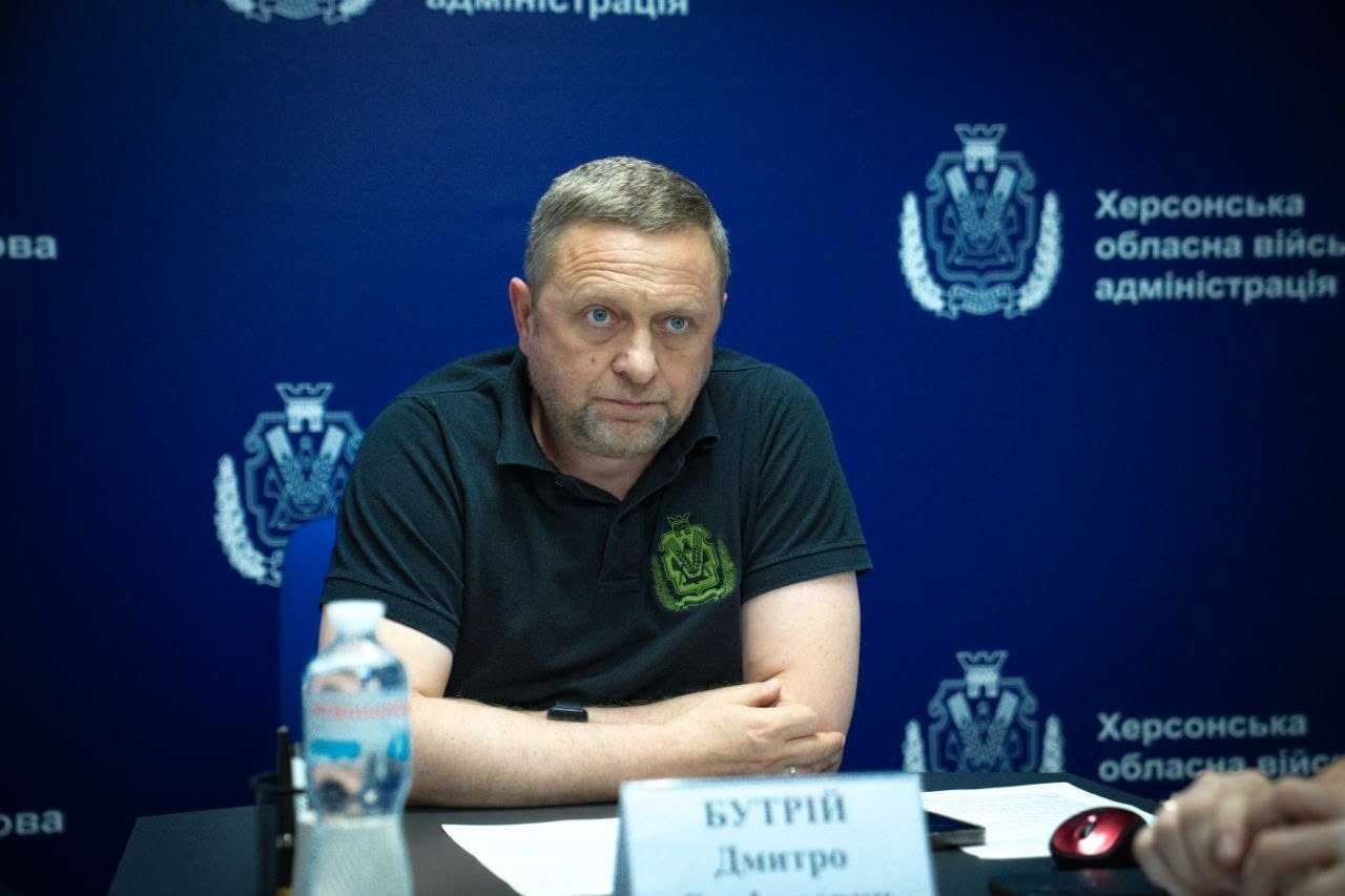 Портретне фото першого заступника начальника Херсонської обласної військової адміністрації Дмитра Бутрія під час онлайн-наради. Чоловік із короткою сивиною та легкою щетиною зображений анфас, він одягнений у чорну футболку-поло з невеликим зеленим гербом на грудях. Він зосереджено дивиться перед собою, склавши руки на столі. Перед ним на столі встановлено табличку з написом «БУТРІЙ Дмитро». На передньому плані ліворуч видно розмиту пластикову пляшку води «Моршинська». Позаду нього розташовано суцільний темно-синій банер із повторюваним білим гербом області та офіційним написом «Херсонська обласна військова адміністрація».