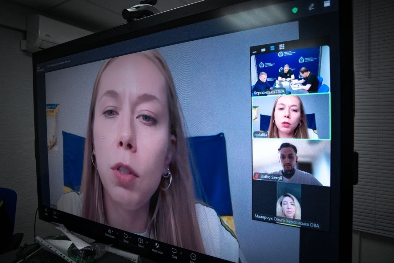 Світлина великого монітора комп'ютера, на якому відображається інтерфейс програми для відеоконференцій під час робочої наради. Головне центральне вікно займає трансляція спікерки — молодої жінки зі світлим волоссям та круглими сережками-кільцями, яка виступає на тлі синьо-жовтого прапора. У правій вертикальній стрічці екрана виведені вікна інших учасників: Верхнє вікно підписане як «Херсонська ОВА», у ньому видно трьох чоловіків за столом у кабінеті. Друге вікно дублює зображення спікерки та підписане «nataliia». Третє вікно транслює чоловіка у сірому одязі з підписом «Boiko Sergii». Нижнє вікно підписане «Малярчук Ольга Херсонська ОВА», у ньому видно учасницю наради.