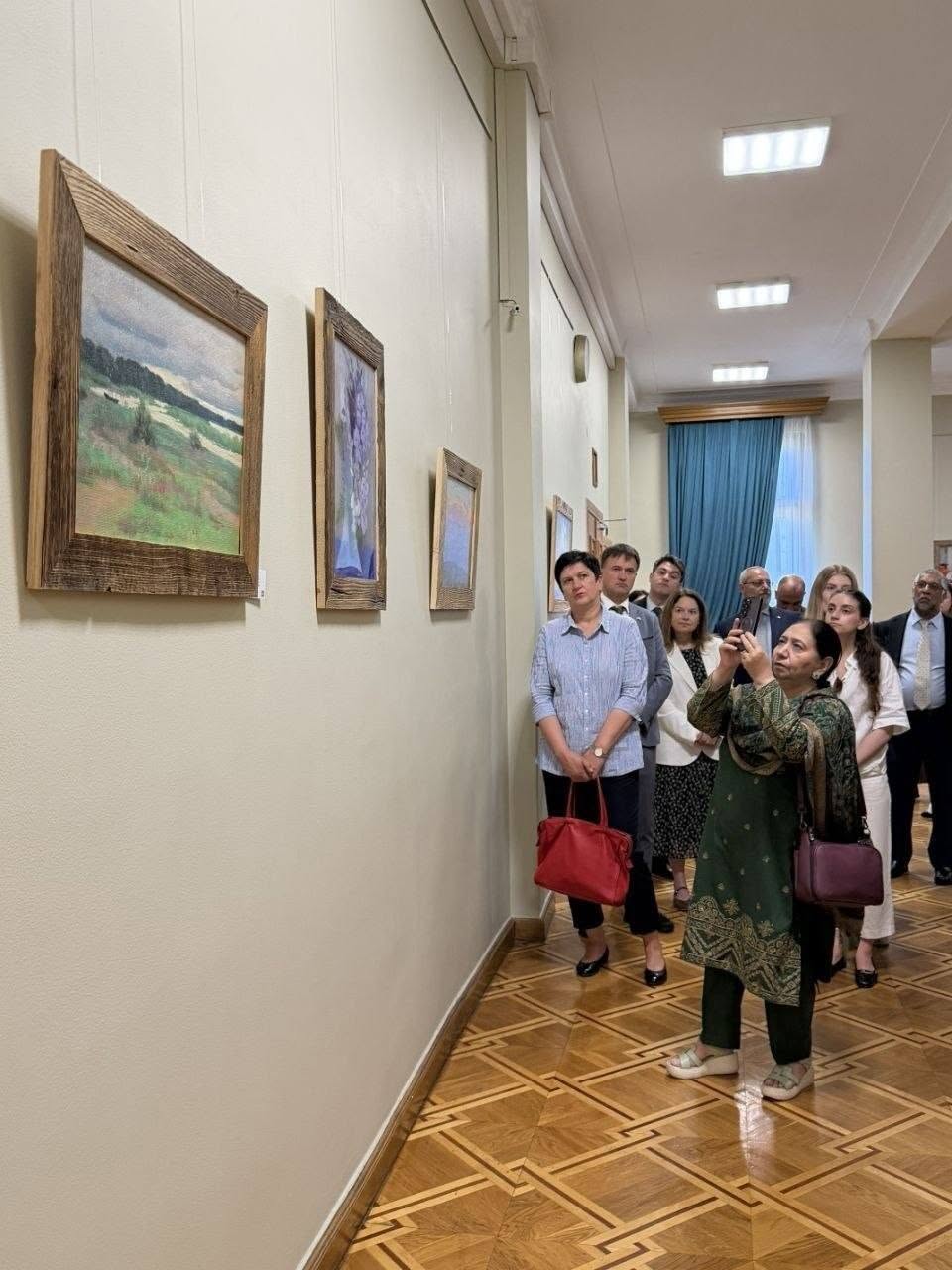 Група відвідувачів та членів іноземної делегації розглядає живописні роботи, розвішані вздовж довгої світлої стіни музейного коридору з лакованим дерев'яним паркетом. На передньому плані праворуч жінка в зеленому національному вбранні (сальвар-каміз) із золотистим візерунком та з темно-бордовою сумкою на плечі фотографує картину на свій смартфон. Поруч із нею та позаду стоять інші екскурсанти, серед яких жінка у блакитній сорочці з червоною сумкою в руках, чоловіки в ділових костюмах та молоді дівчата. На стелі залу горить ряд квадратних вбудованих ламп, а в глибині коридору видно біле вікно з великою синьою шторою.