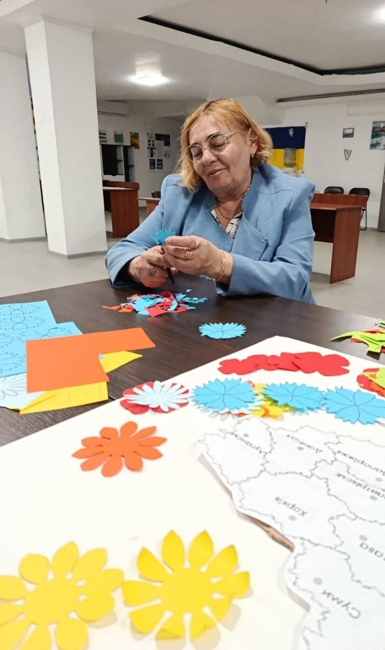Процес створення патріотичного колажу. Великим планом на передньому плані зображено білий аркуш із надрукованою картою України, де підписані назви обласних центрів, зокрема «Харків», «Суми». Поруч із мапою лежать вирізані деталі паперових квітів: жовті соняшники та блакитні волошки.  На задньому плані за столом сидить жінка в окулярах та світло-блакитному піджаку поверх блузки. Вона посміхається і тримає в руках ножиці, збираючи докупи елементи паперової квітки блакитного кольору. На столі перед нею розкидані дрібні кольорові обрізки паперу.