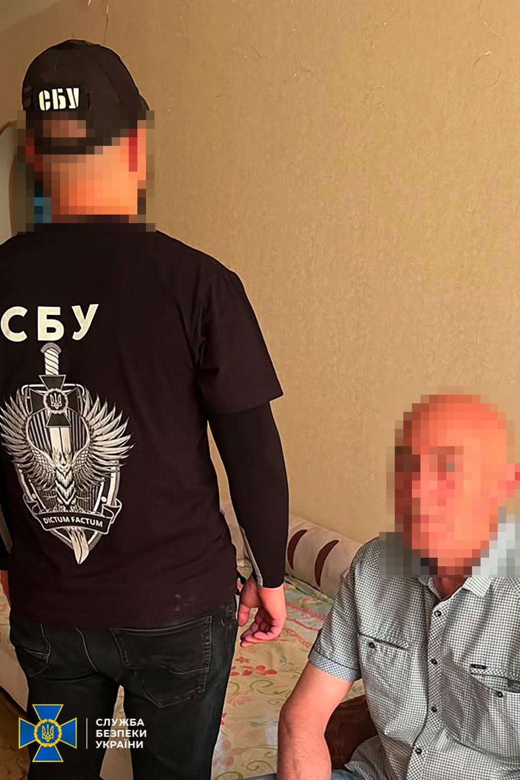 Співробітник СБУ проводить слідчі дії з підозрюваним у житловій кімнаті. Ліворуч спиною до камери стоїть оперативник у чорній кепці з білим написом «СБУ» та чорній футболці, на спині якої нанесено великий білий бренд-логотип відомства: меч вістрям донизу, накладений на щит із козацьким хрестом, крилами та супроводжувальним девізом на стрічці: «DICTUM FACTUM». Його ліва рука одягнена в чорний довгий тактичний рукав.  Праворуч на дивані з візерунчастим покривалом сидить літній лисий чоловік у сірій сорочці в дрібну клітинку з коротким рукавом. Голова та обличчя підозрюваного повністю приховані великим цифровим розмиттям (пікселями). Тло за ними — однотонна бежева стіна.